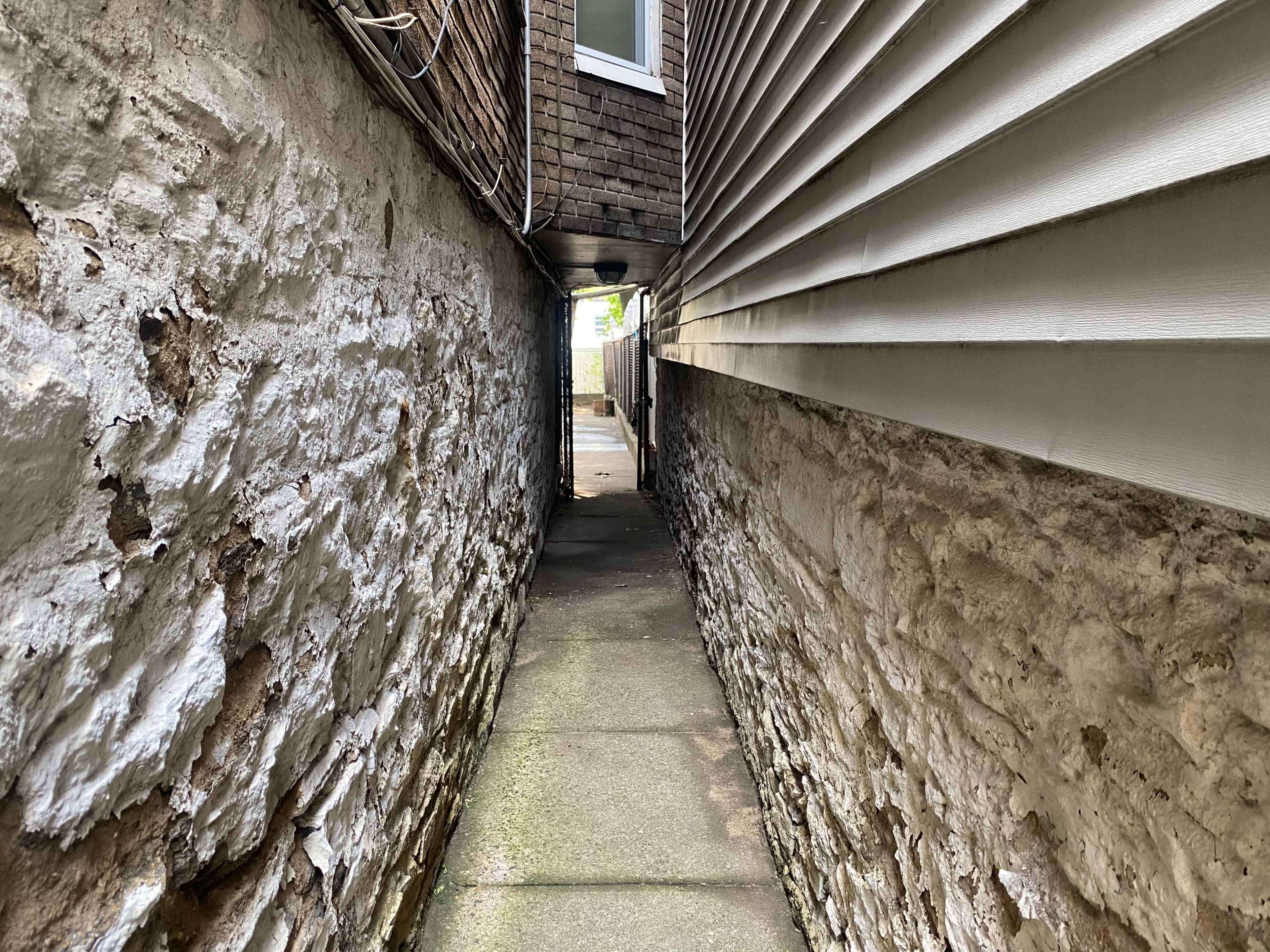 A narrow alleyway between two buildings, with stone walls on one side and a siding surface on the other.
