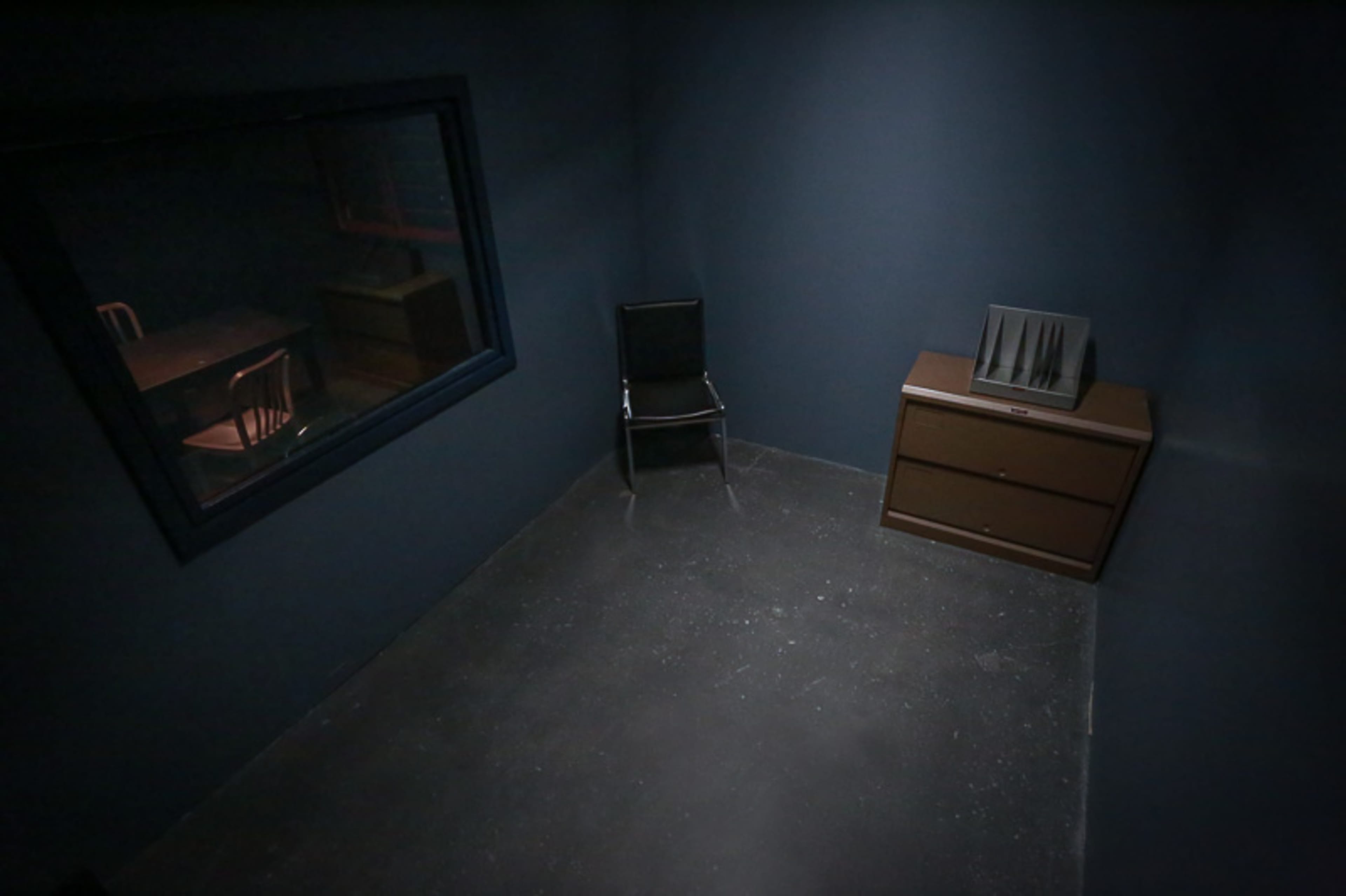 A dimly lit, empty room with a chair, a dresser, and a window looking into another space.