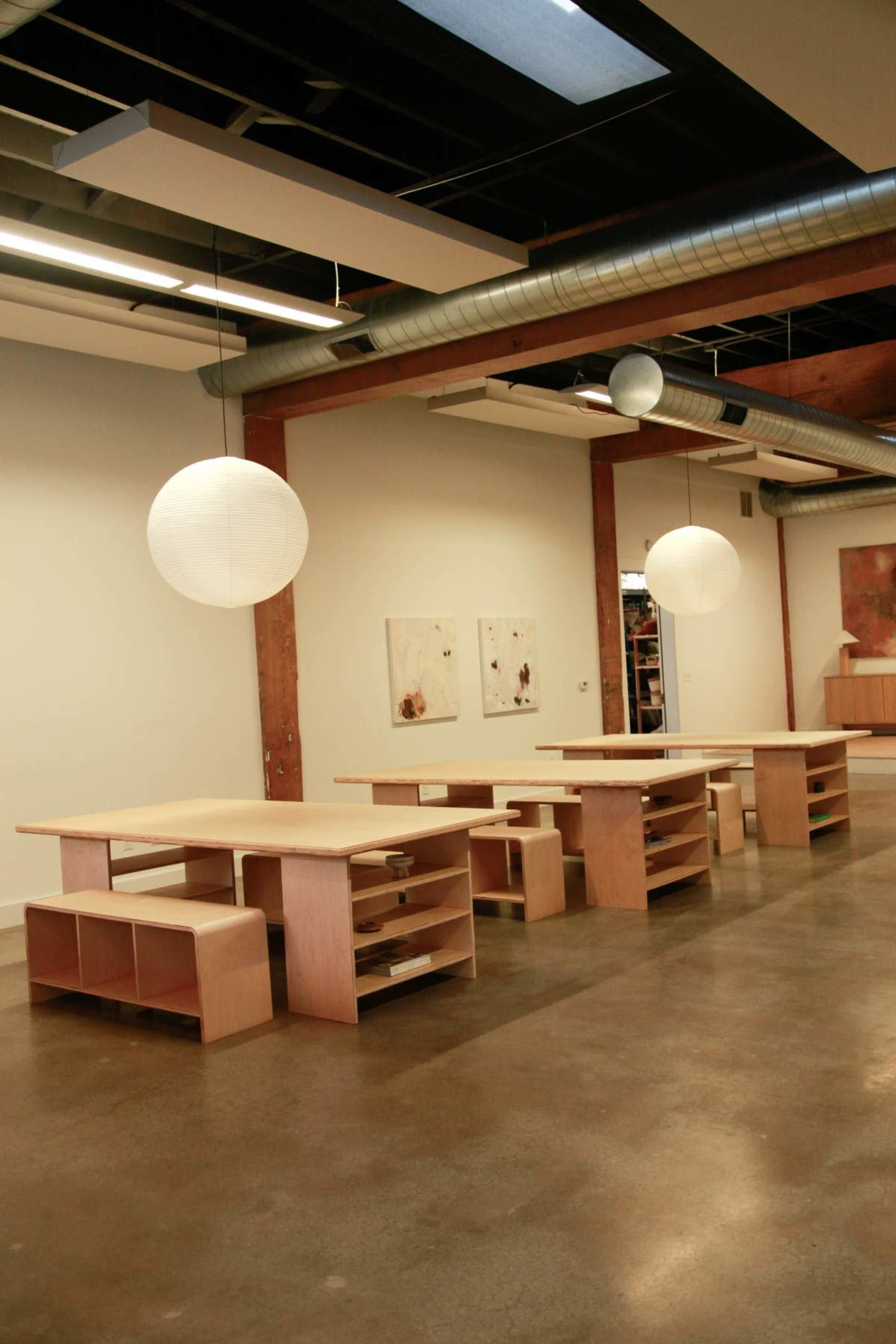 The image shows a spacious, modern room with several wooden tables arranged in a row, hanging pendant lights, and artwork on the walls.