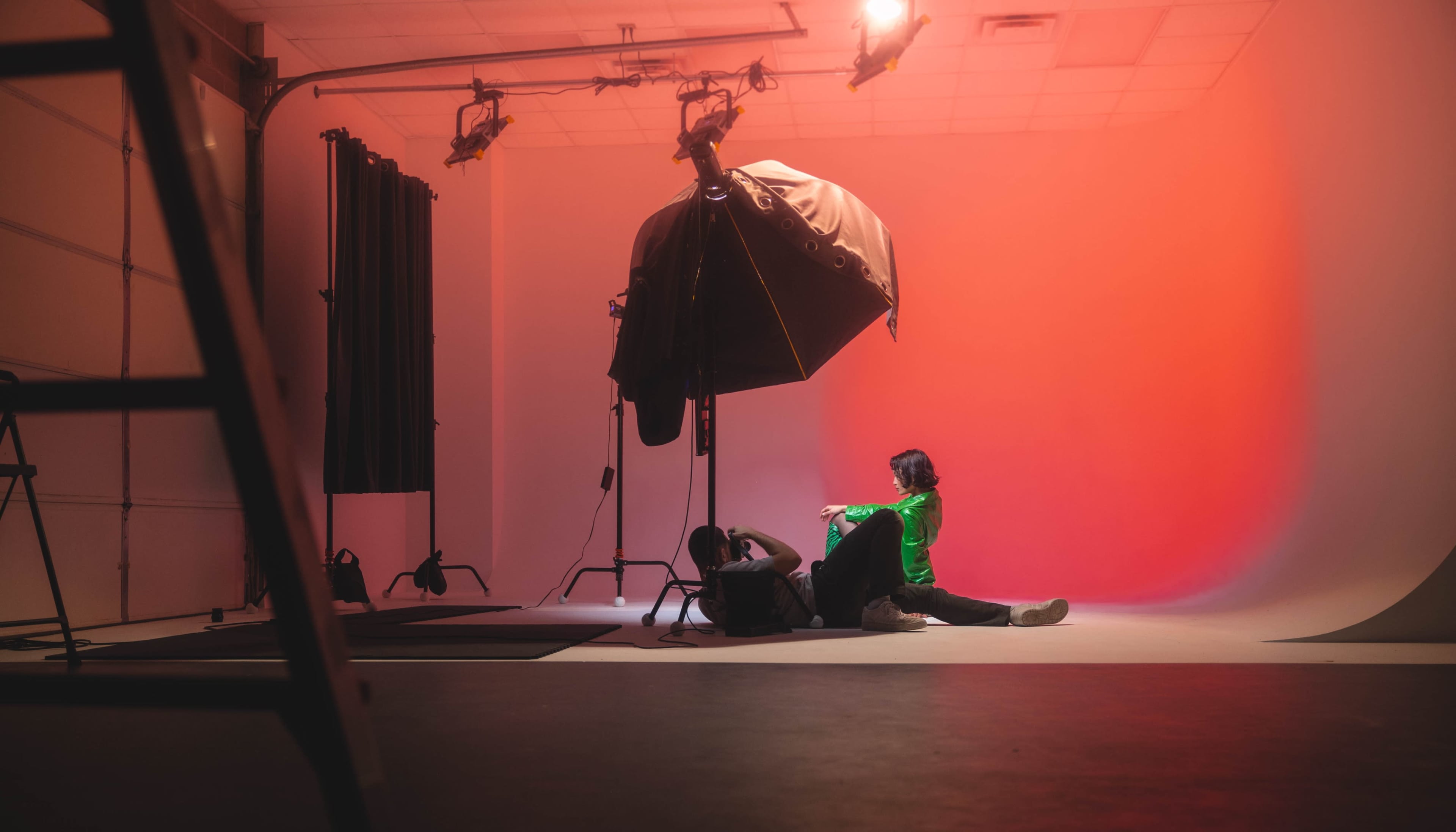 A person wearing a green jacket is seated on the floor of a photography studio with a large softbox lighting setup and a gradient backdrop.