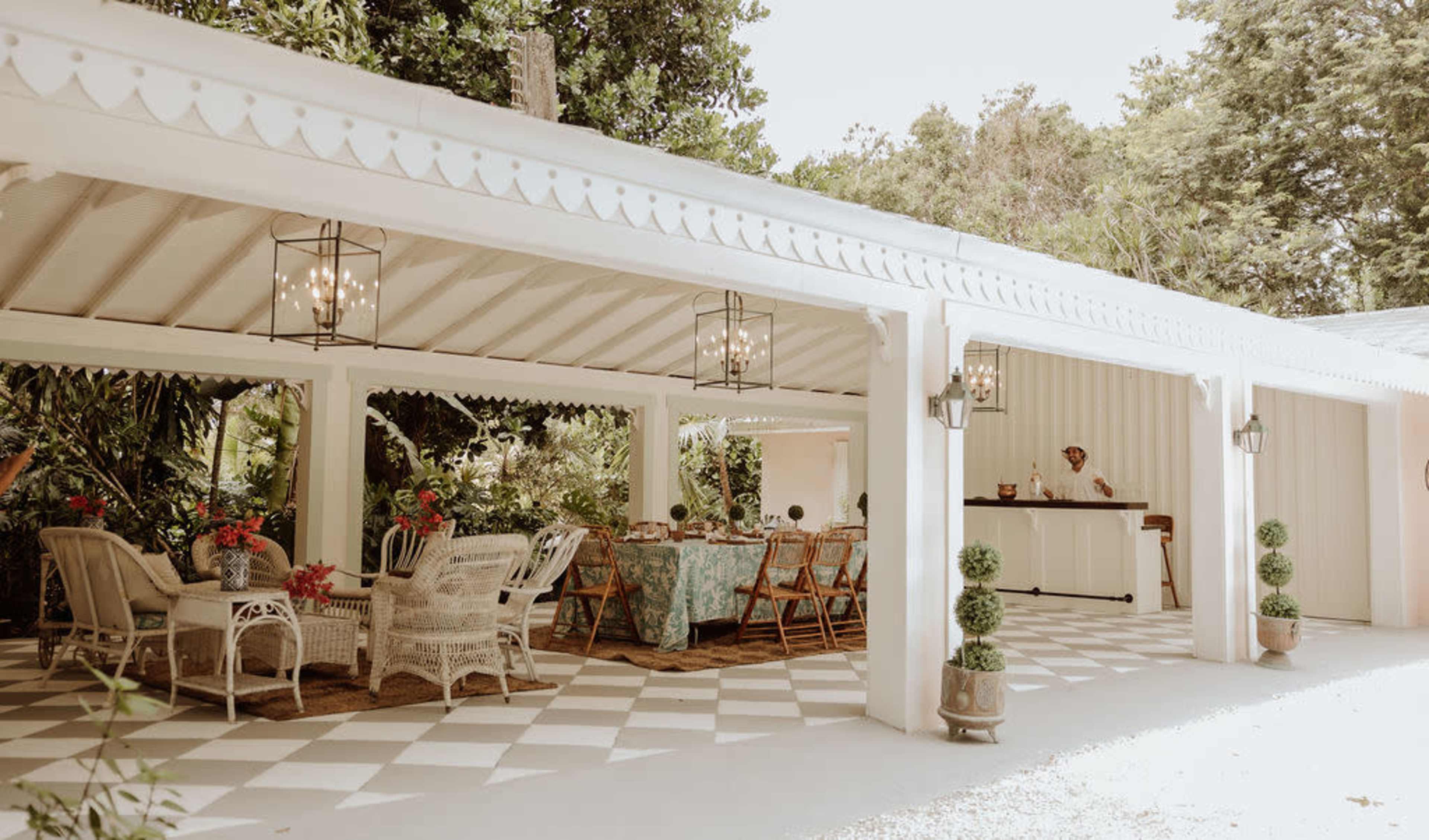 The image shows a covered outdoor dining area with wicker furniture, a checkered floor, and tables set for a meal surrounded by plants.