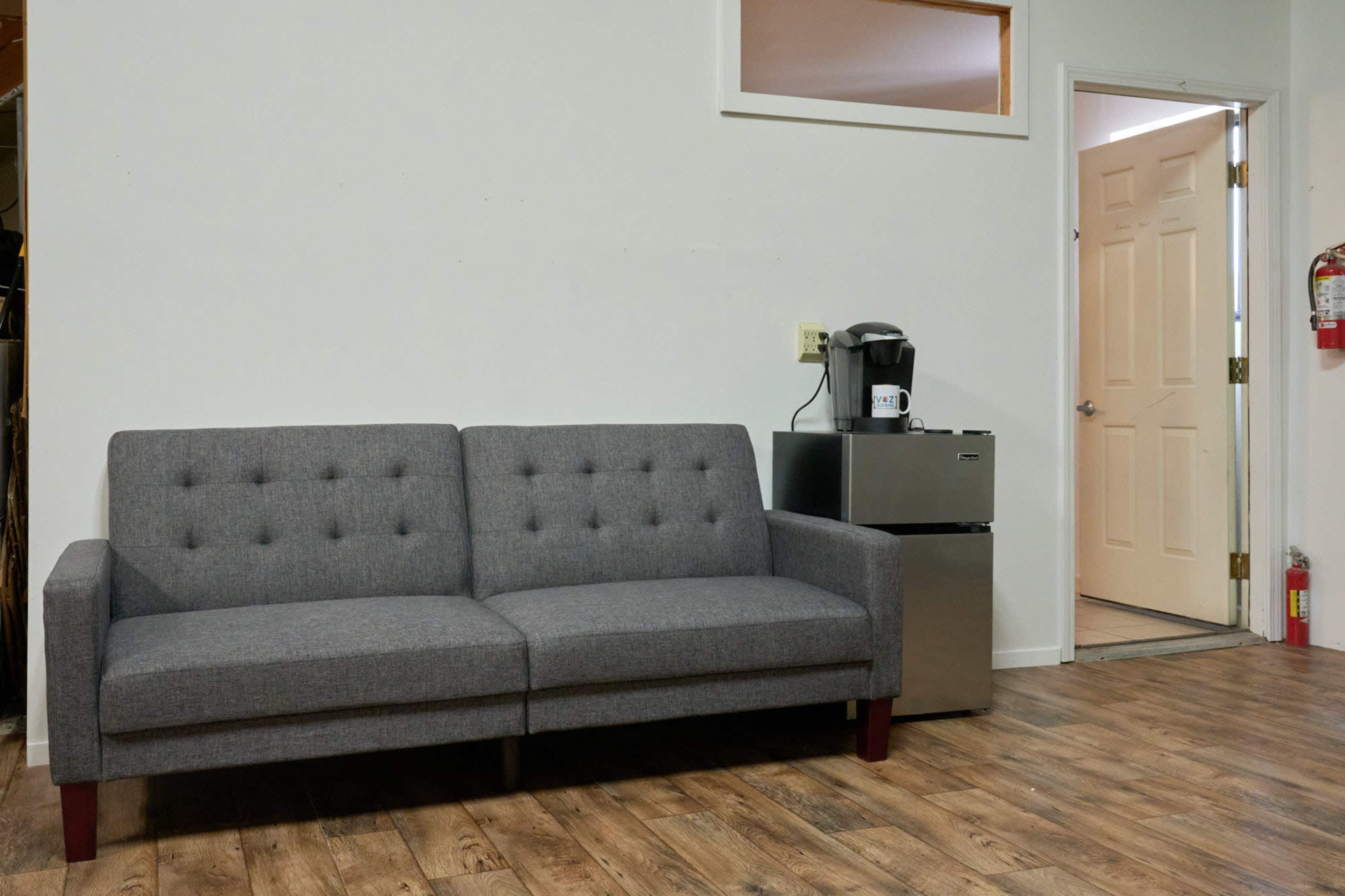 A gray upholstered sofa facing a small kitchen area with a coffee maker and a closed door to another room.