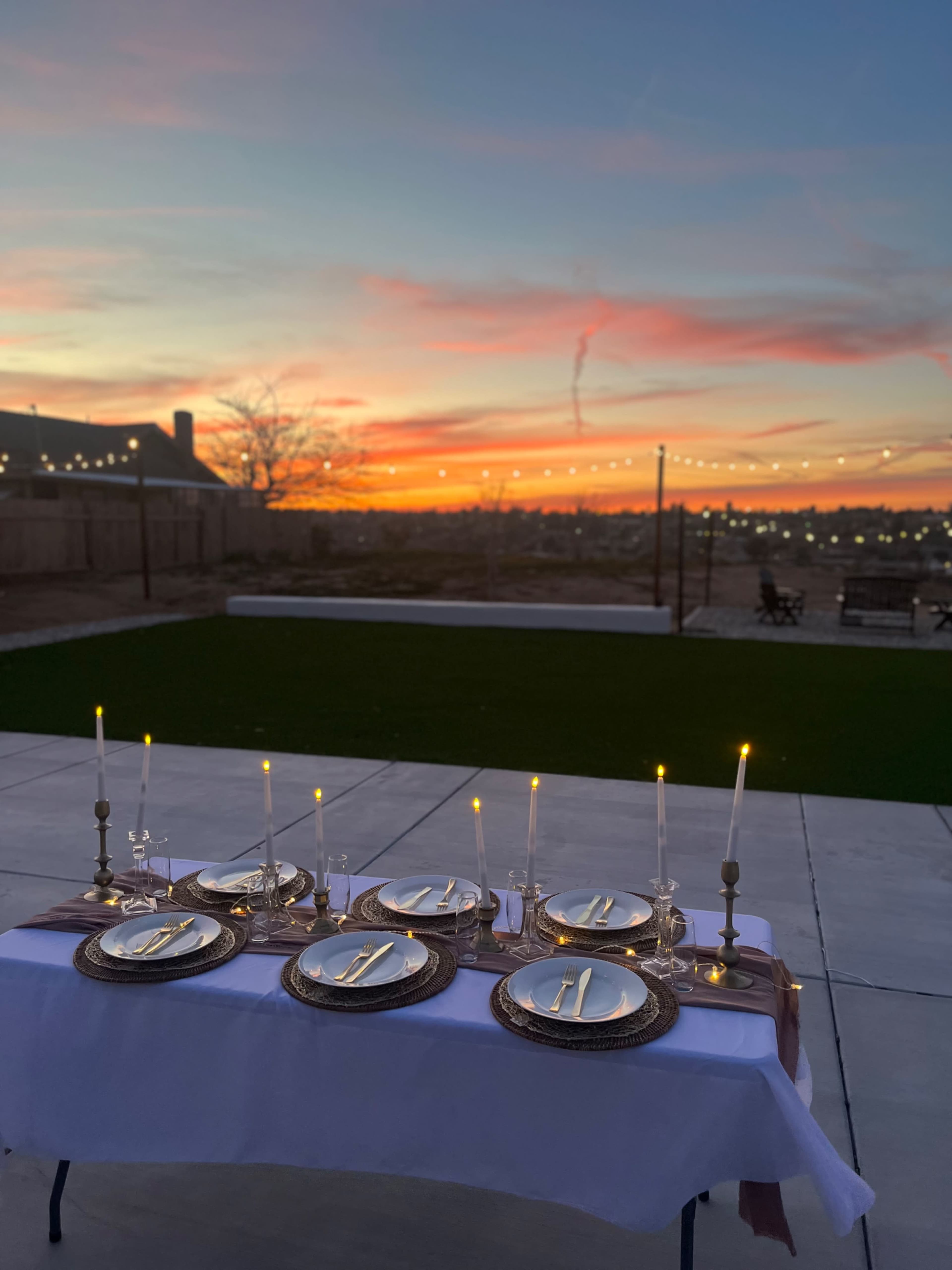 A neatly set outdoor dining table with plates, cutlery, and candles is positioned against a vibrant sunset sky.