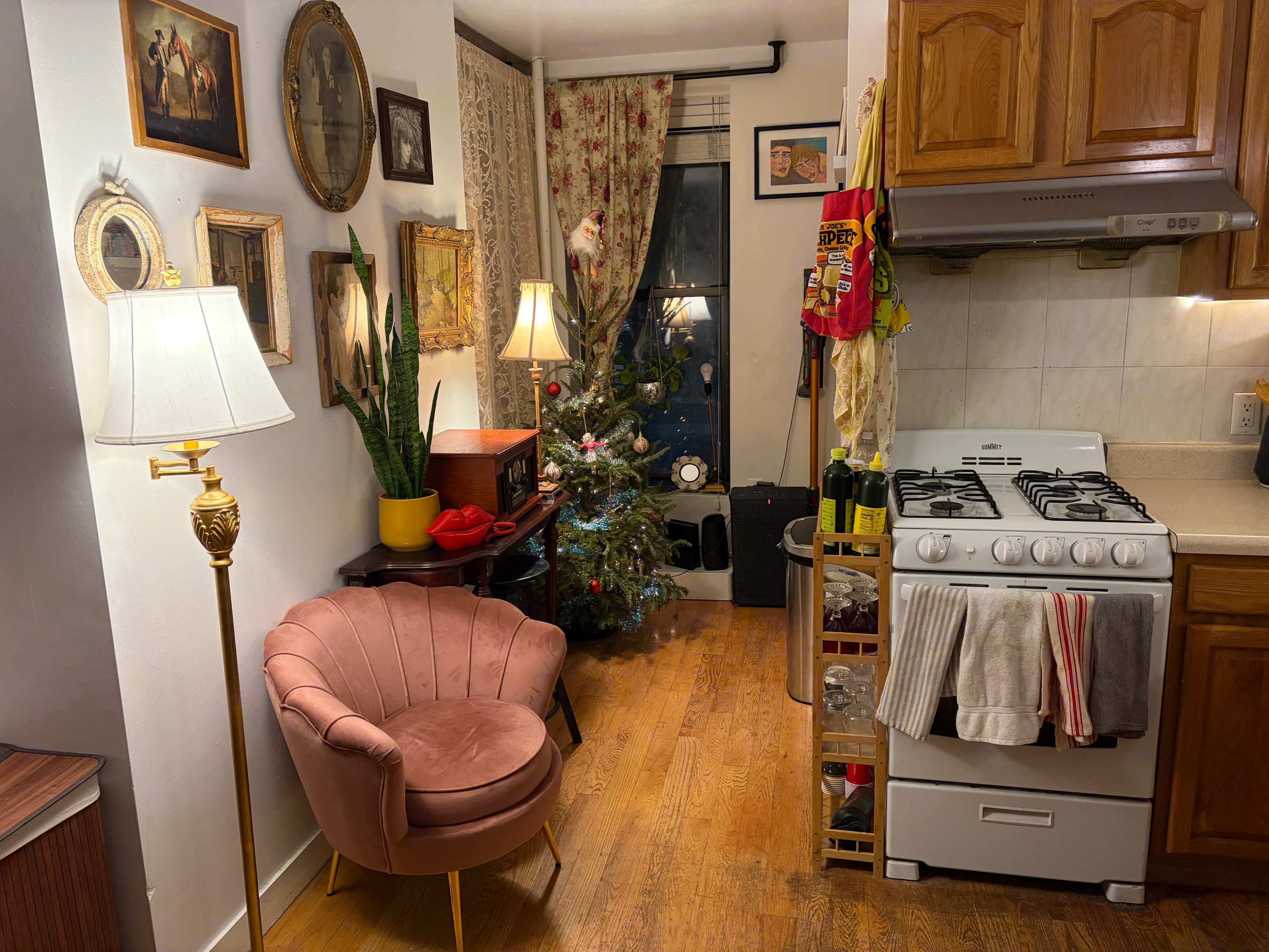 The image shows a cozy kitchen area with a gas stove, wooden cabinets, and a small Christmas tree beside a pink armchair, adorned with decorative wall frames and a lamp.