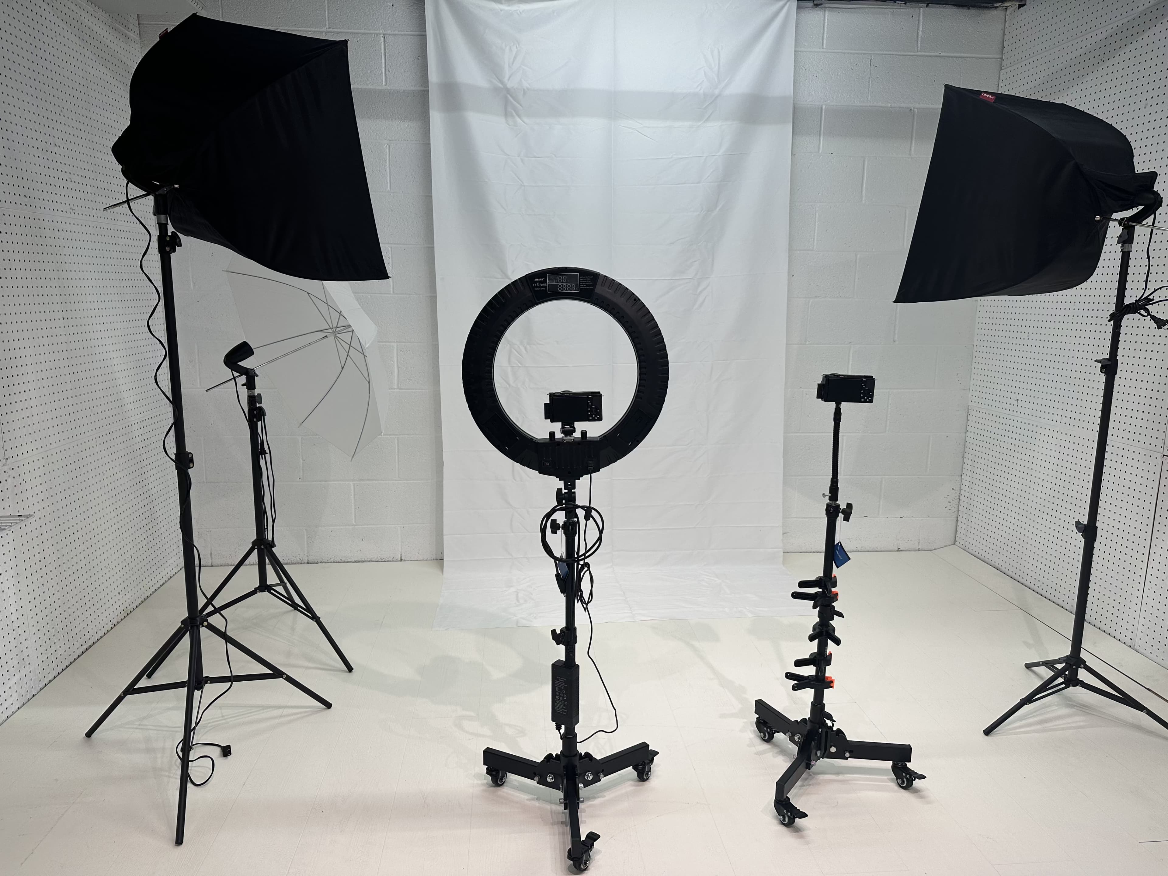A photography studio setup with three lighting stands and a circular ring light positioned in front of a white backdrop.