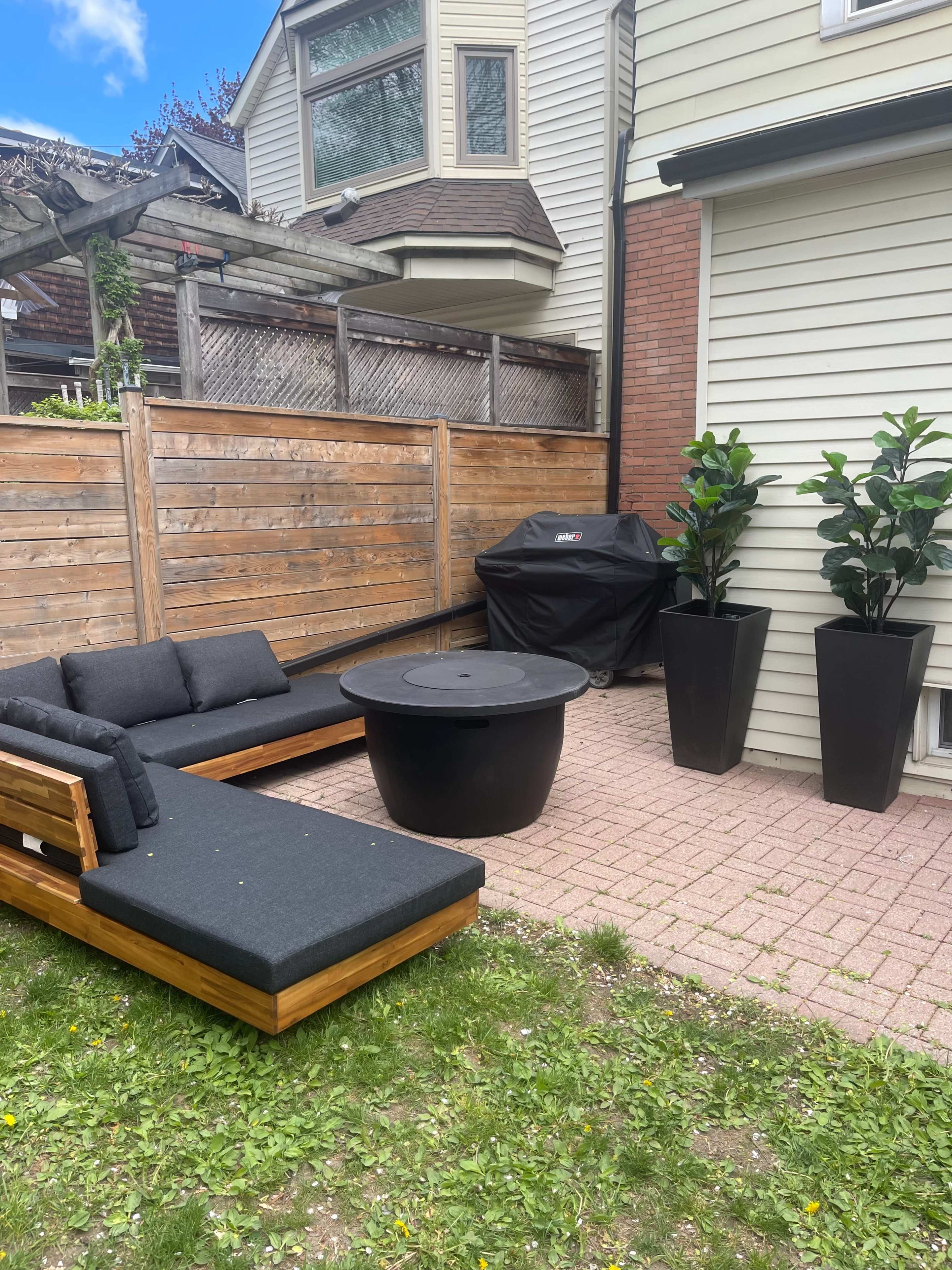 A patio area with a wooden seating arrangement, a round black table, a grill, and tall planters with plants, enclosed by a wooden fence.