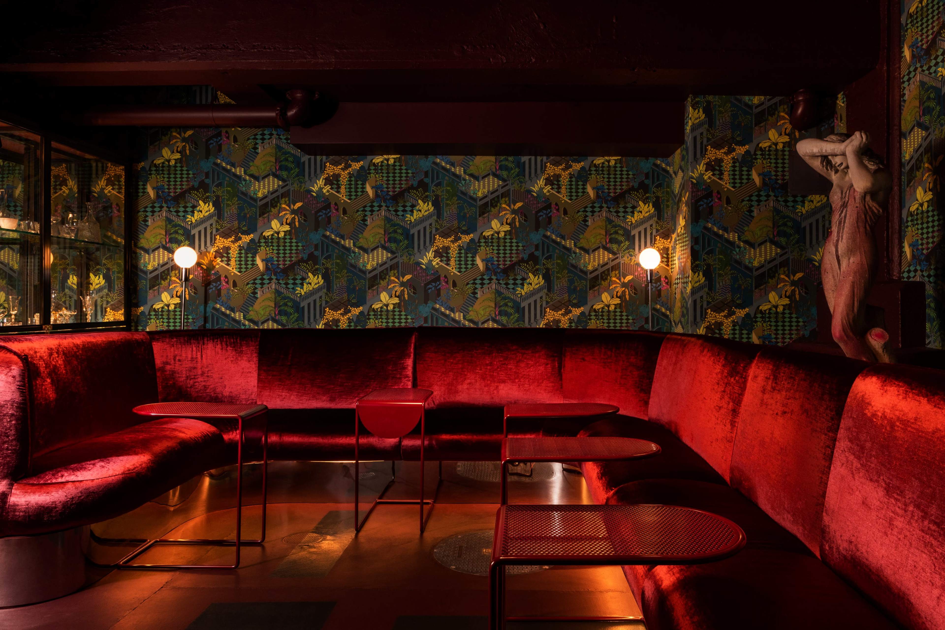 The image shows a dimly lit lounge area with a red velvet curved sofa, small circular tables, and patterned wallpaper featuring geometric designs.