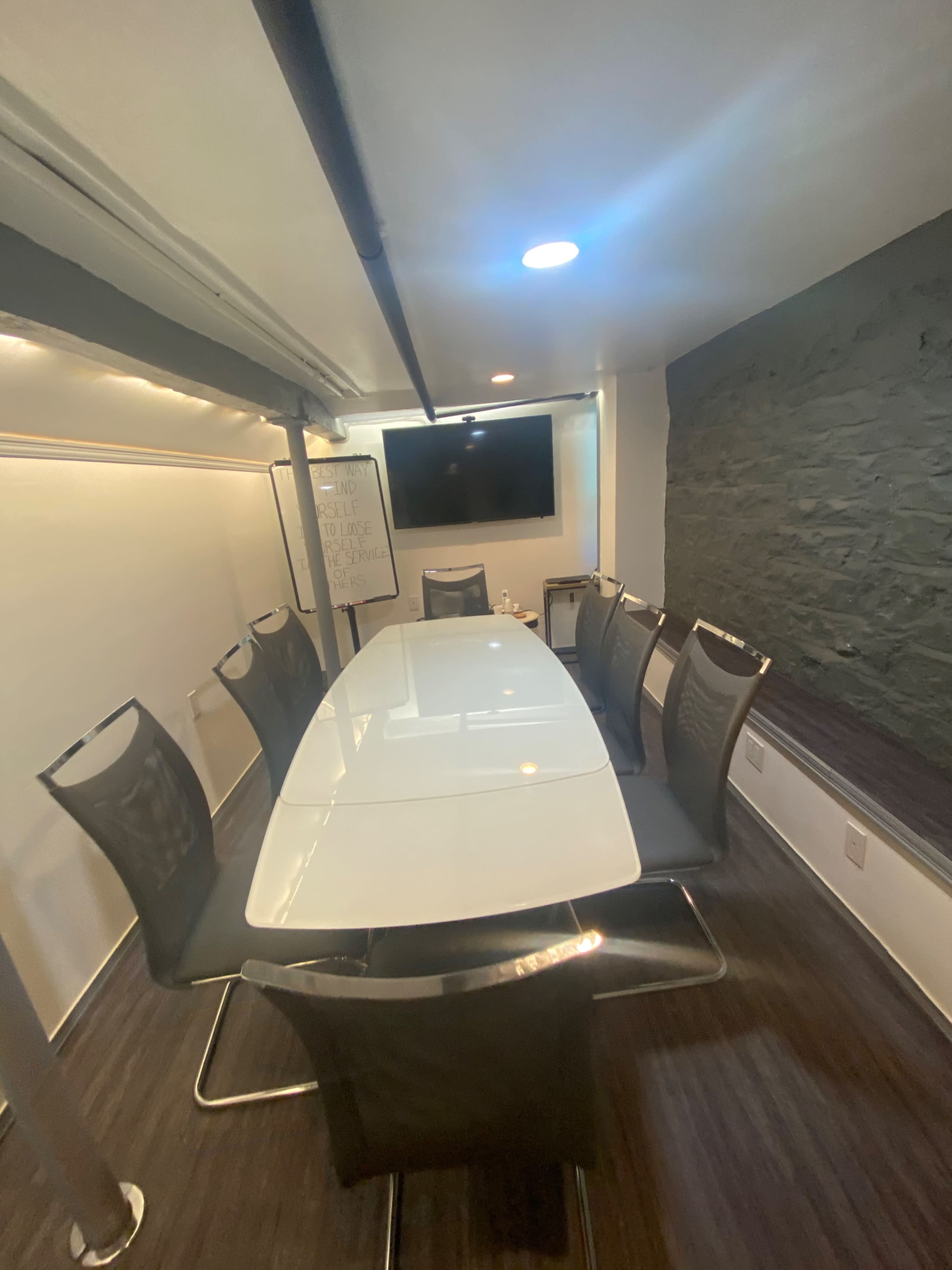 A modern conference room features a large white table surrounded by eight chairs, with a wall-mounted screen and a flip chart in the background.