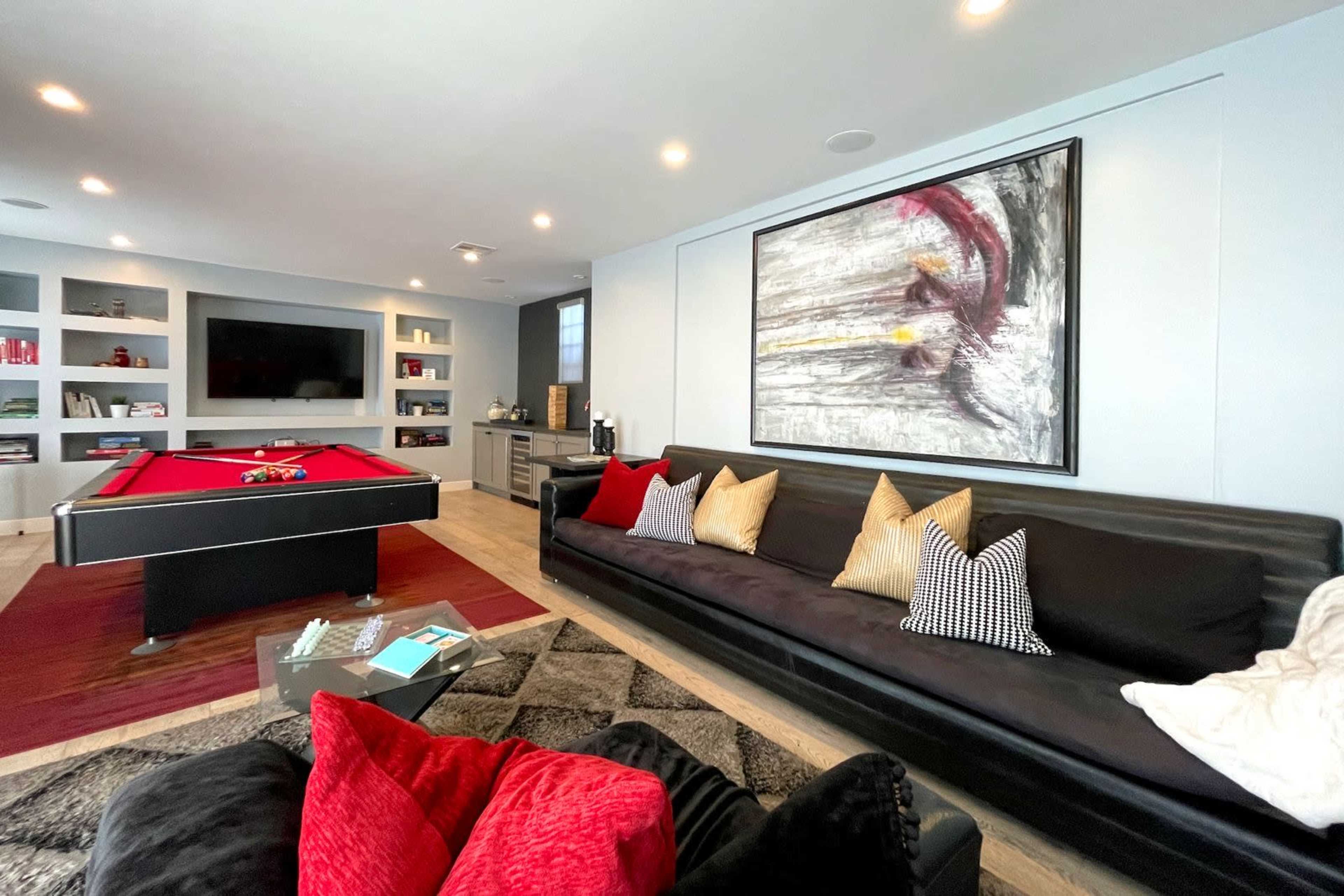 The image shows a modern game room featuring a billiards table, a black sofa with decorative pillows, and a large wall-mounted television surrounded by shelves.