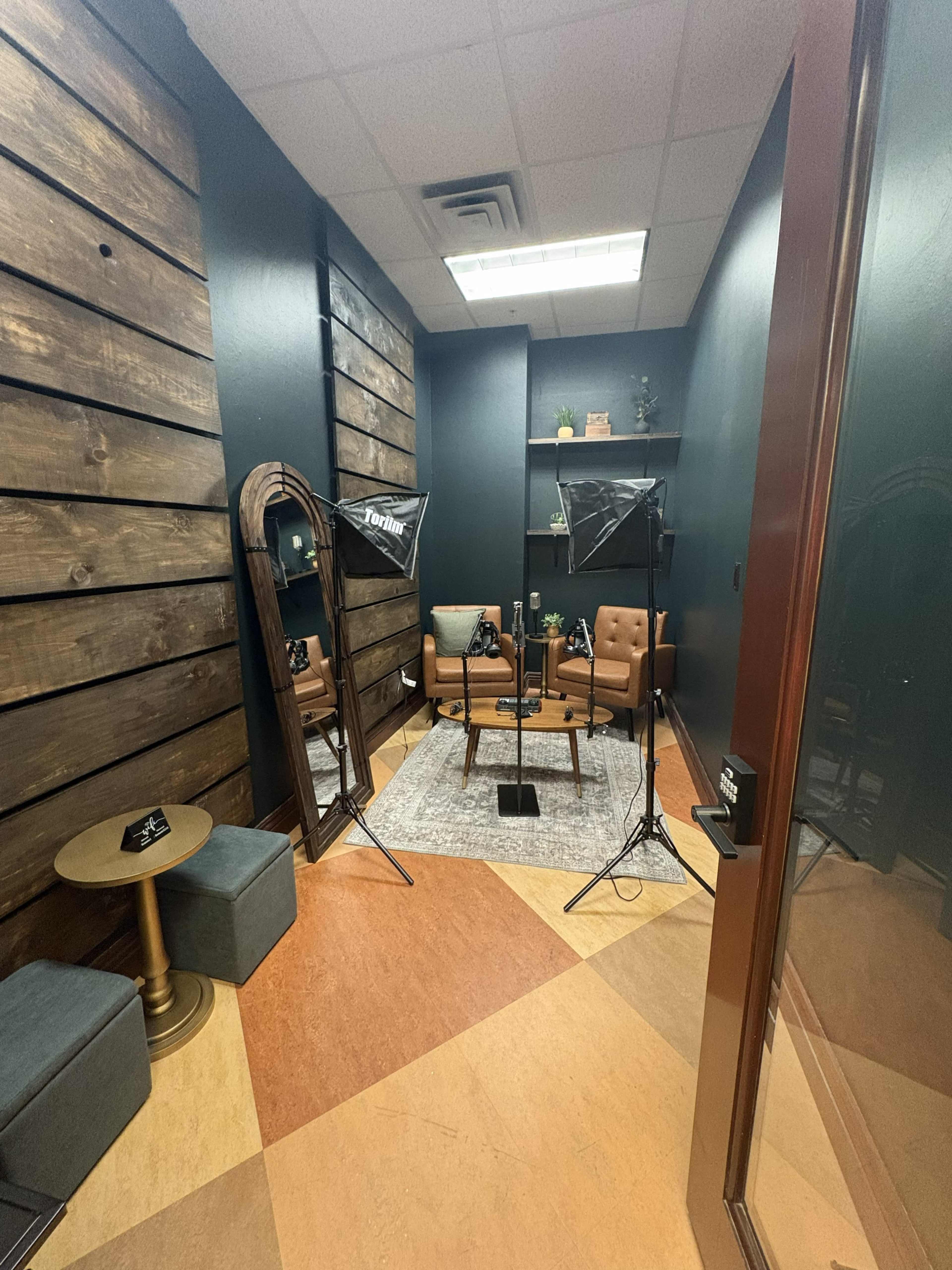 The image shows a small room featuring a seating area with two chairs, a mirror, and lighting equipment, against a backdrop of textured wooden walls.