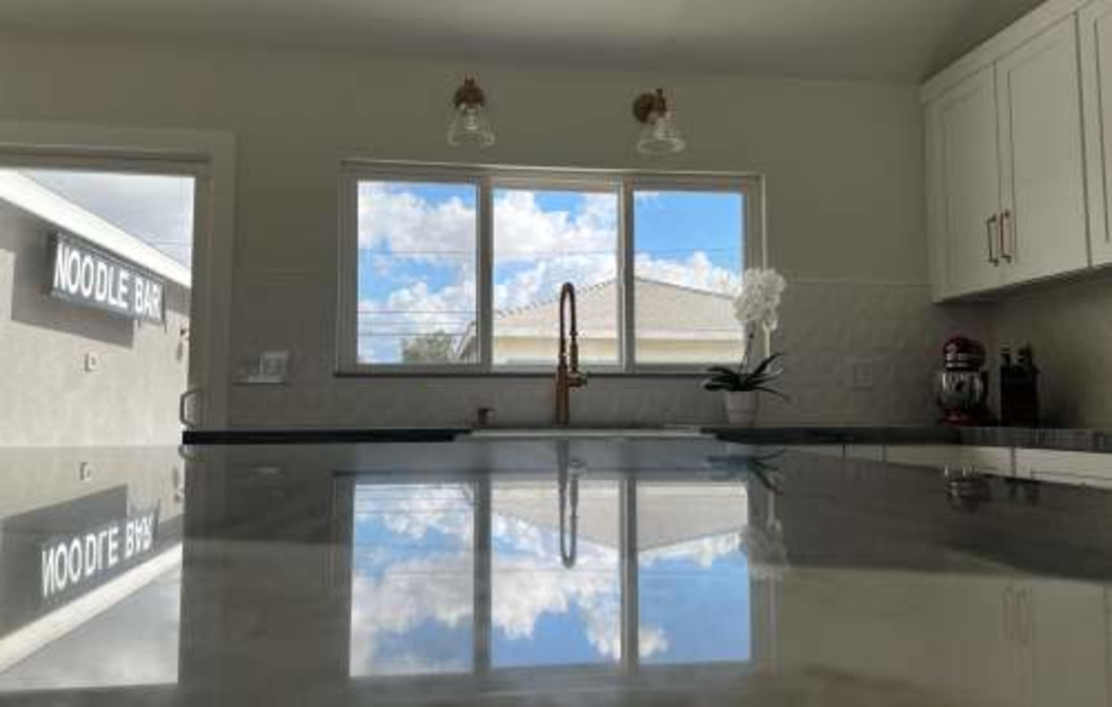 The image shows a modern kitchen with a marble countertop, a window reflecting clouds, and a nearby sign that reads "NOODLE BAR."
