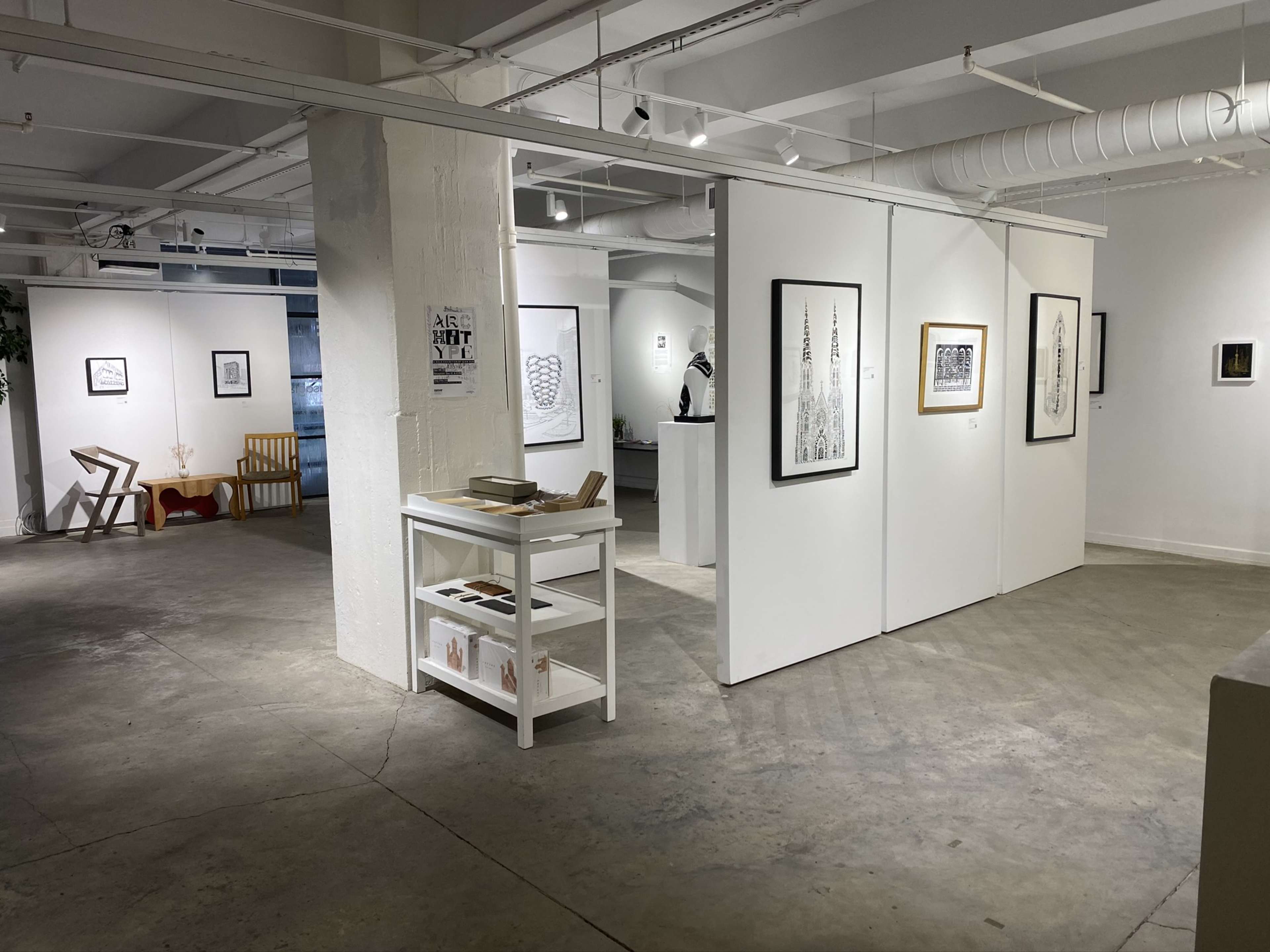 An art gallery with white walls displaying framed artworks and a small white cart holding books.