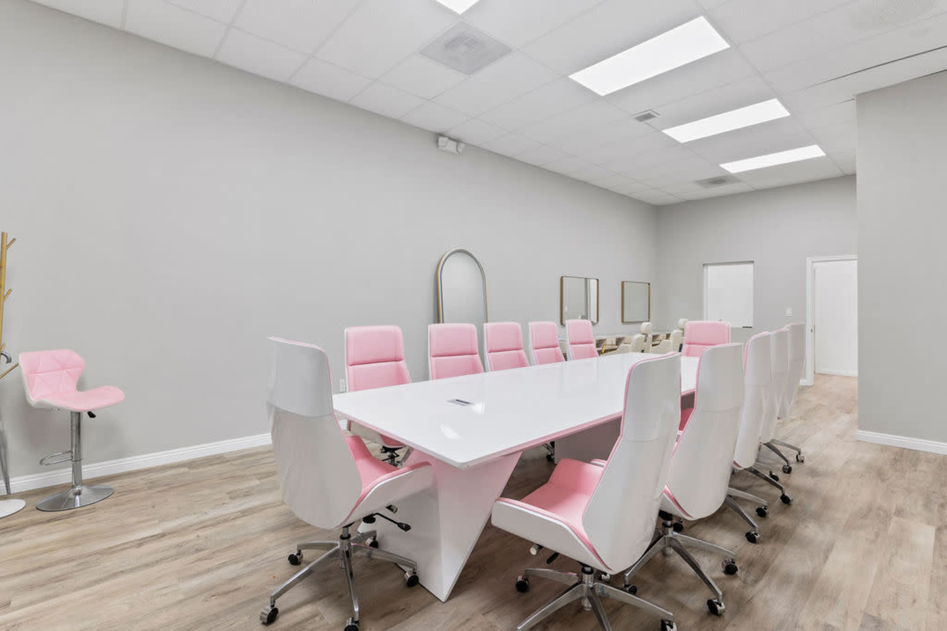 A modern conference room features a large white table surrounded by pink and white chairs, with large windows and minimalist decor.