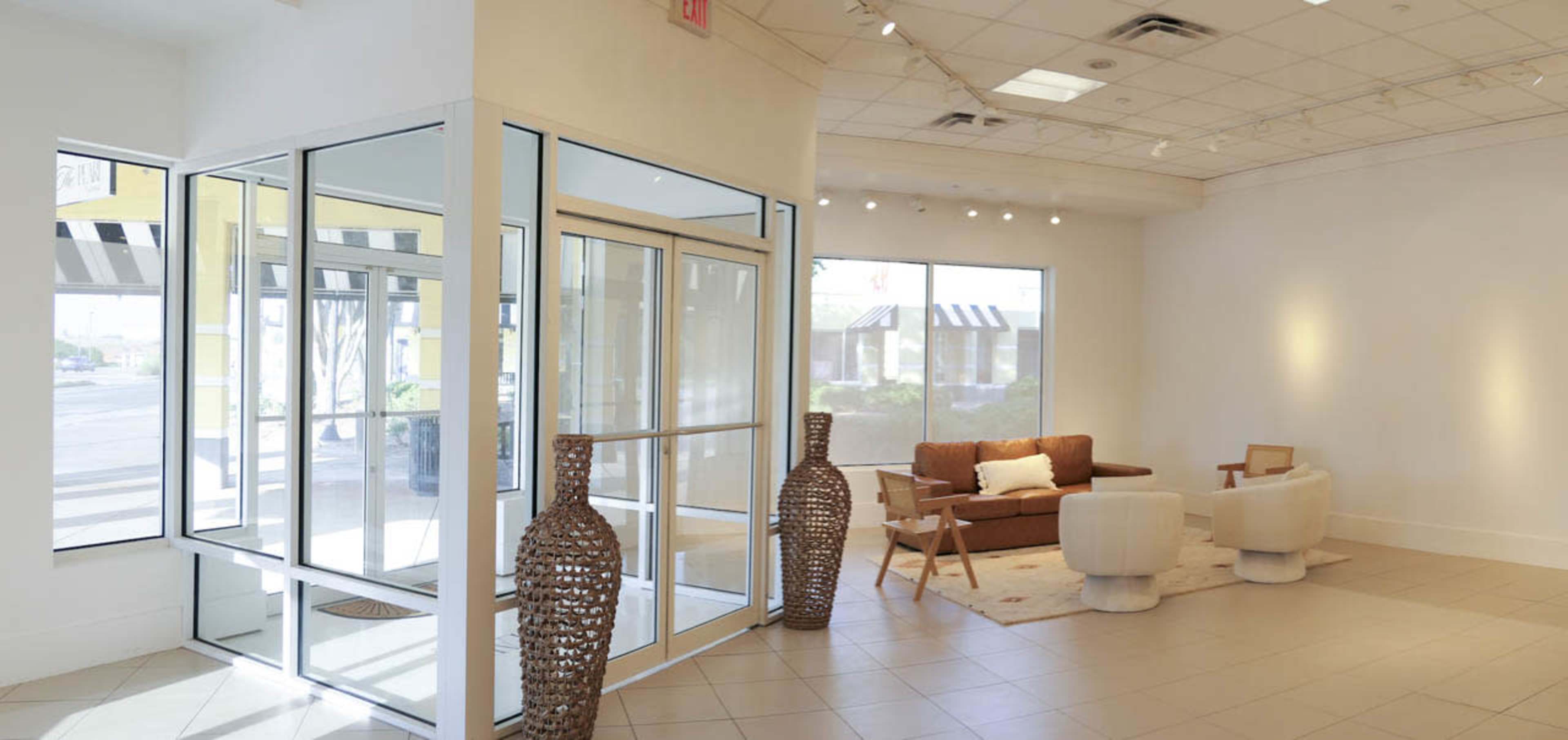 The image shows a modern, spacious lobby area featuring a glass entryway, a brown sofa, white chairs, and tall decorative vases.