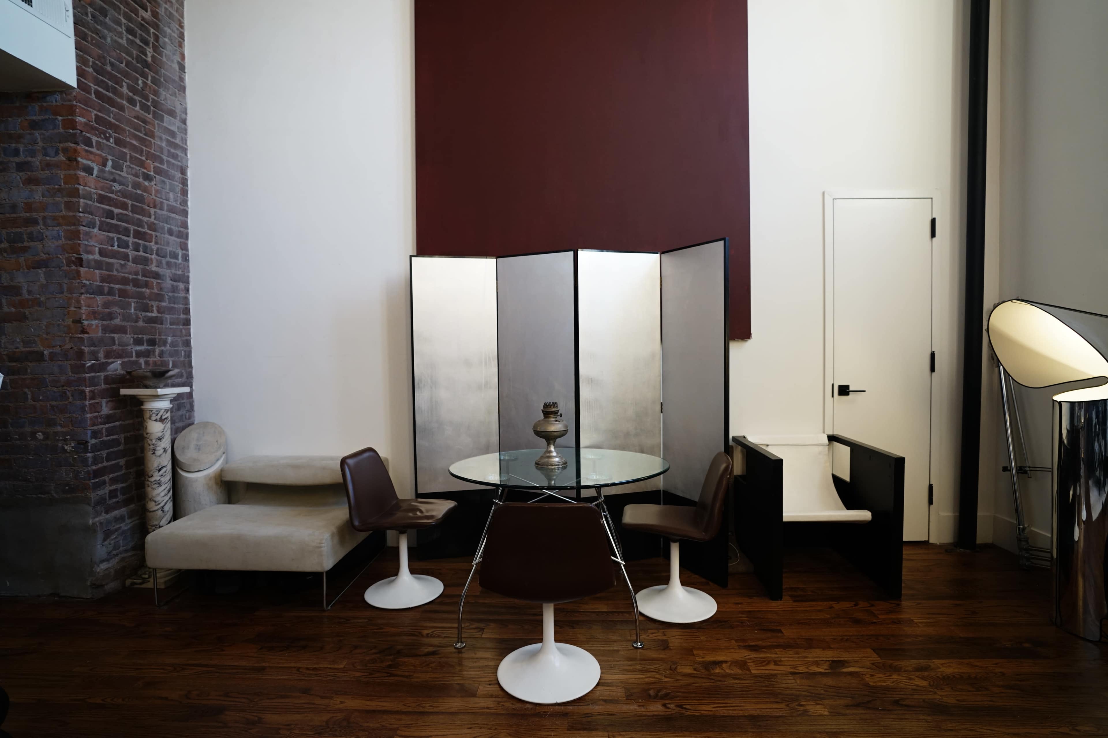 The scene features a modern dining area with a round glass table, four chairs, a folding screen, and various seating styles against a backdrop of exposed brick and a large section of painted wall.