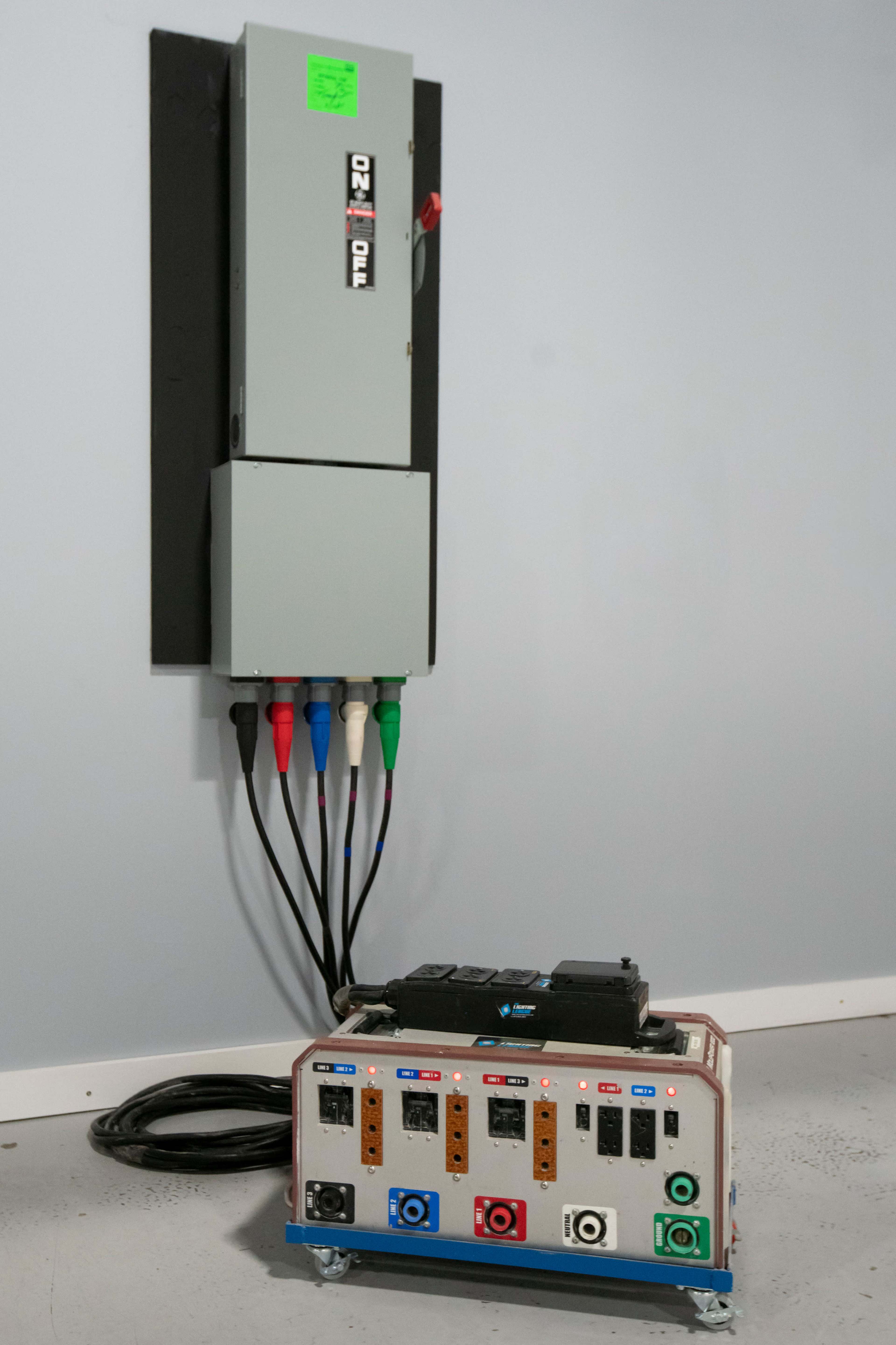 An electrical panel mounted on a wall above a control box with various colored connectors and buttons on a floor surface.