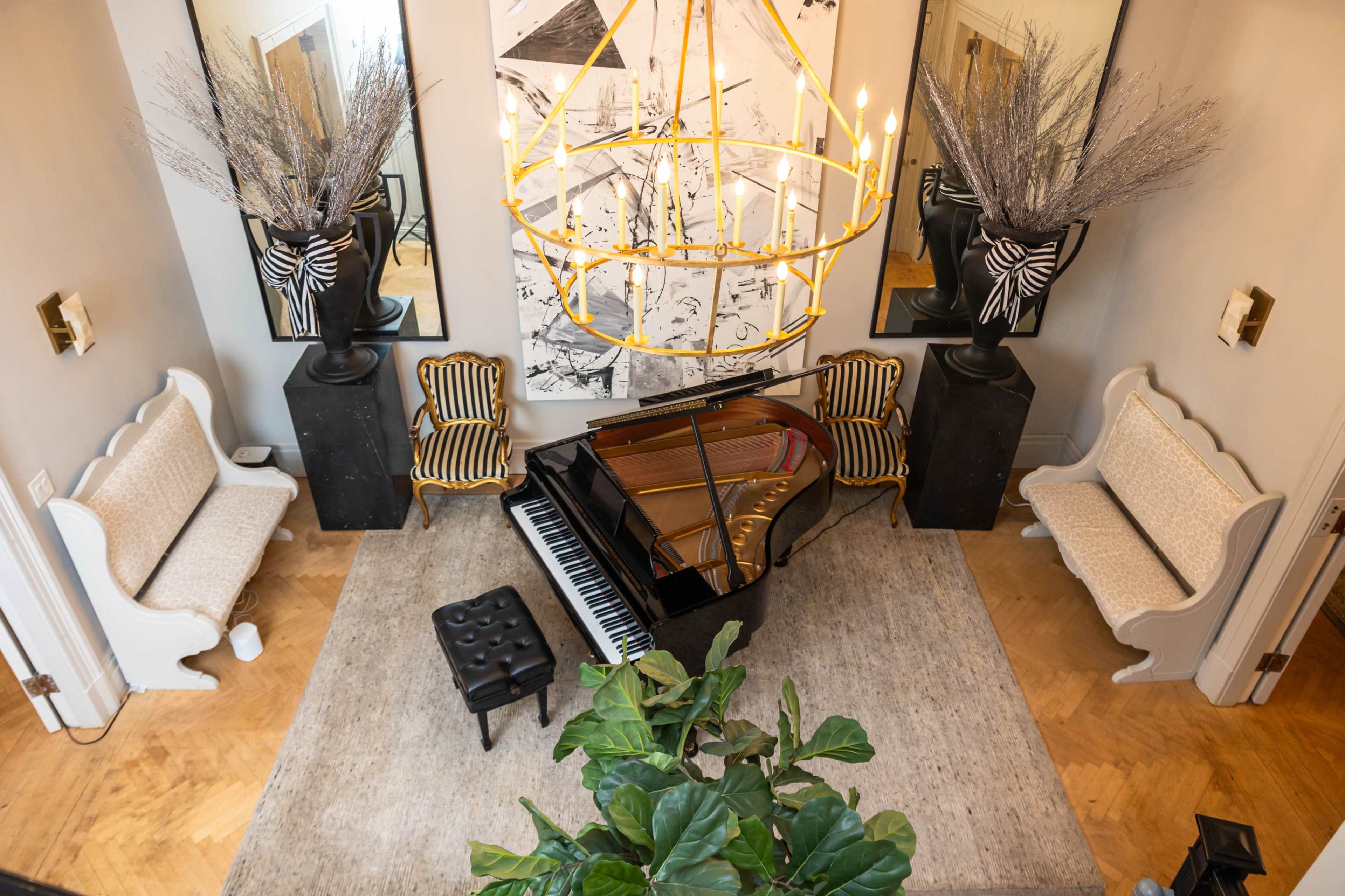 The image shows a living room with a grand piano at the center, flanked by two patterned chairs and surrounded by decorative plants and large mirrors.