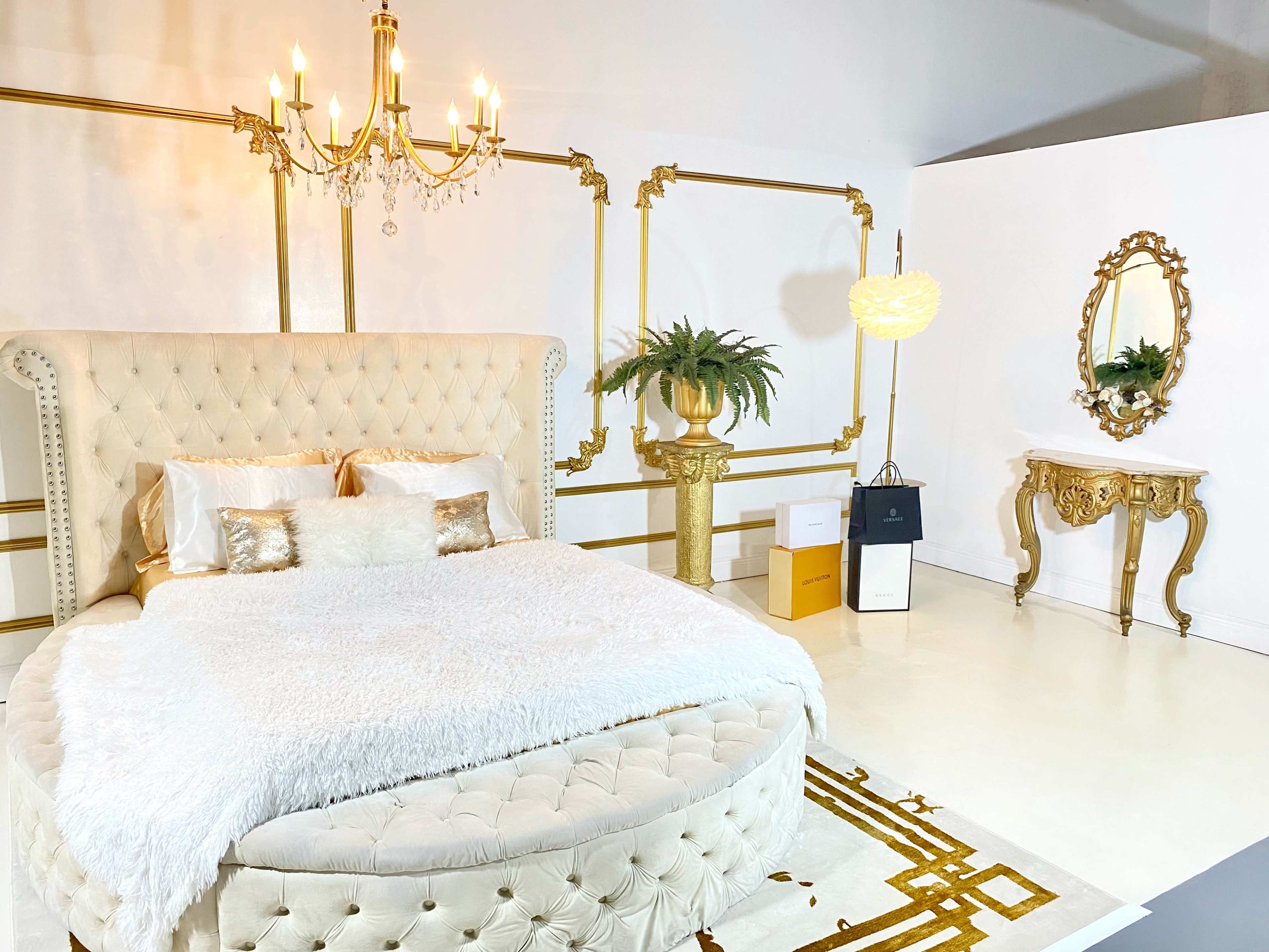 An elegant bedroom featuring a round upholstered bed with a plush white blanket, a chandelier overhead, and decorative gold accents on the walls and furniture.