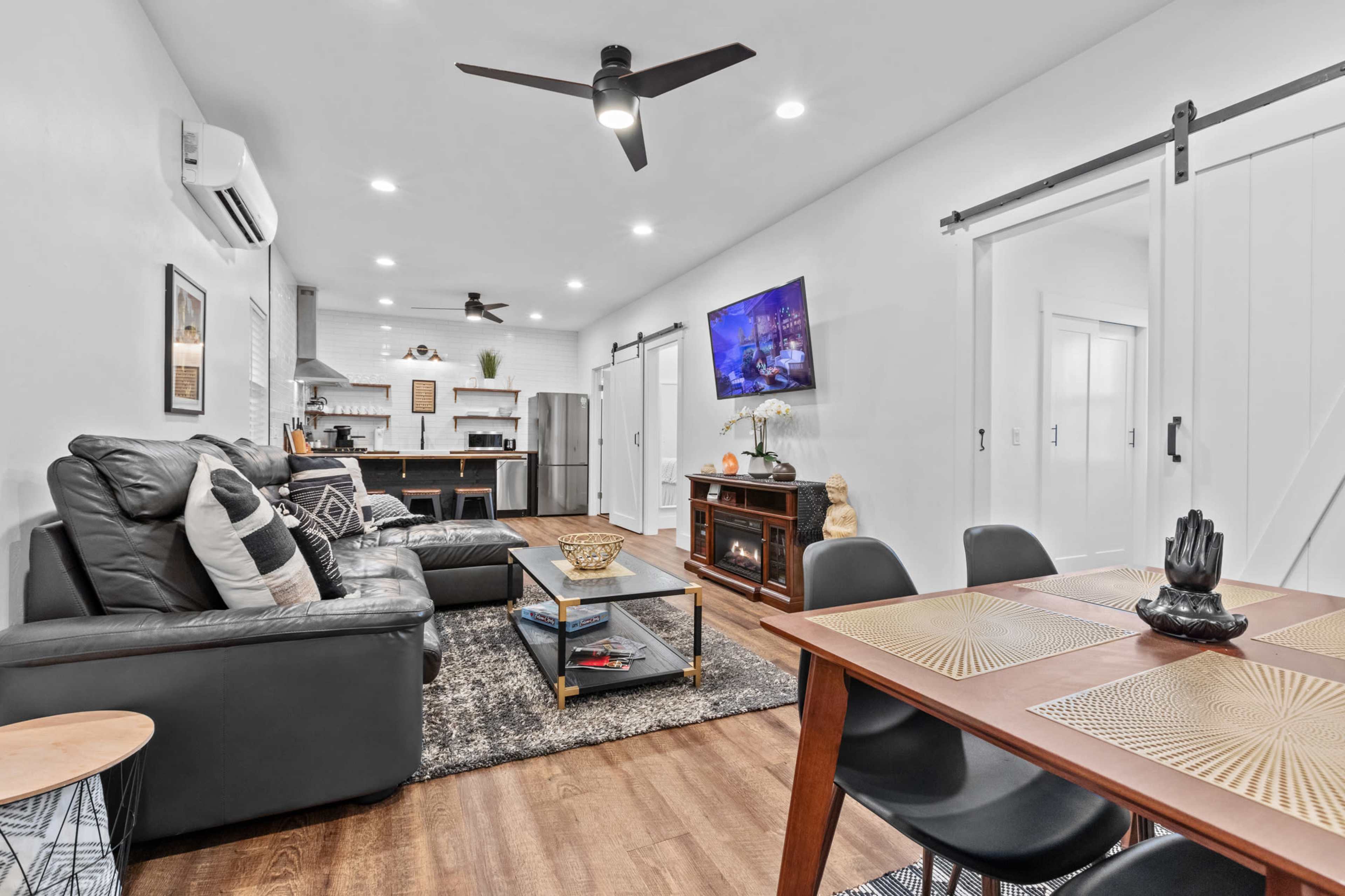 The image shows a modern living space featuring a black leather sofa, a dining table, and a kitchen in the background with stainless steel appliances.