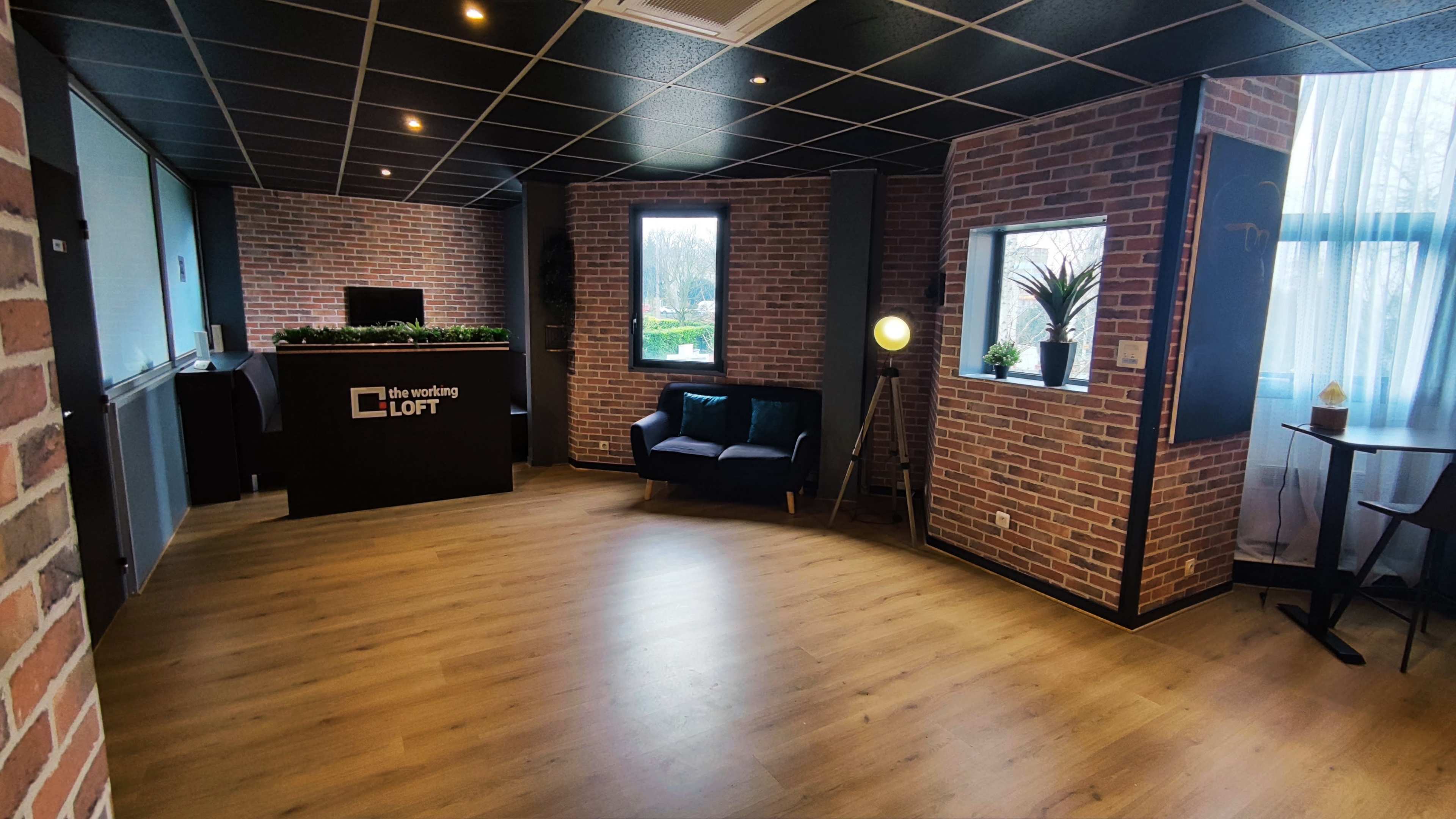A modern office reception area featuring a brick wall, wooden flooring, a seating area, and a small desk with decorative plants.