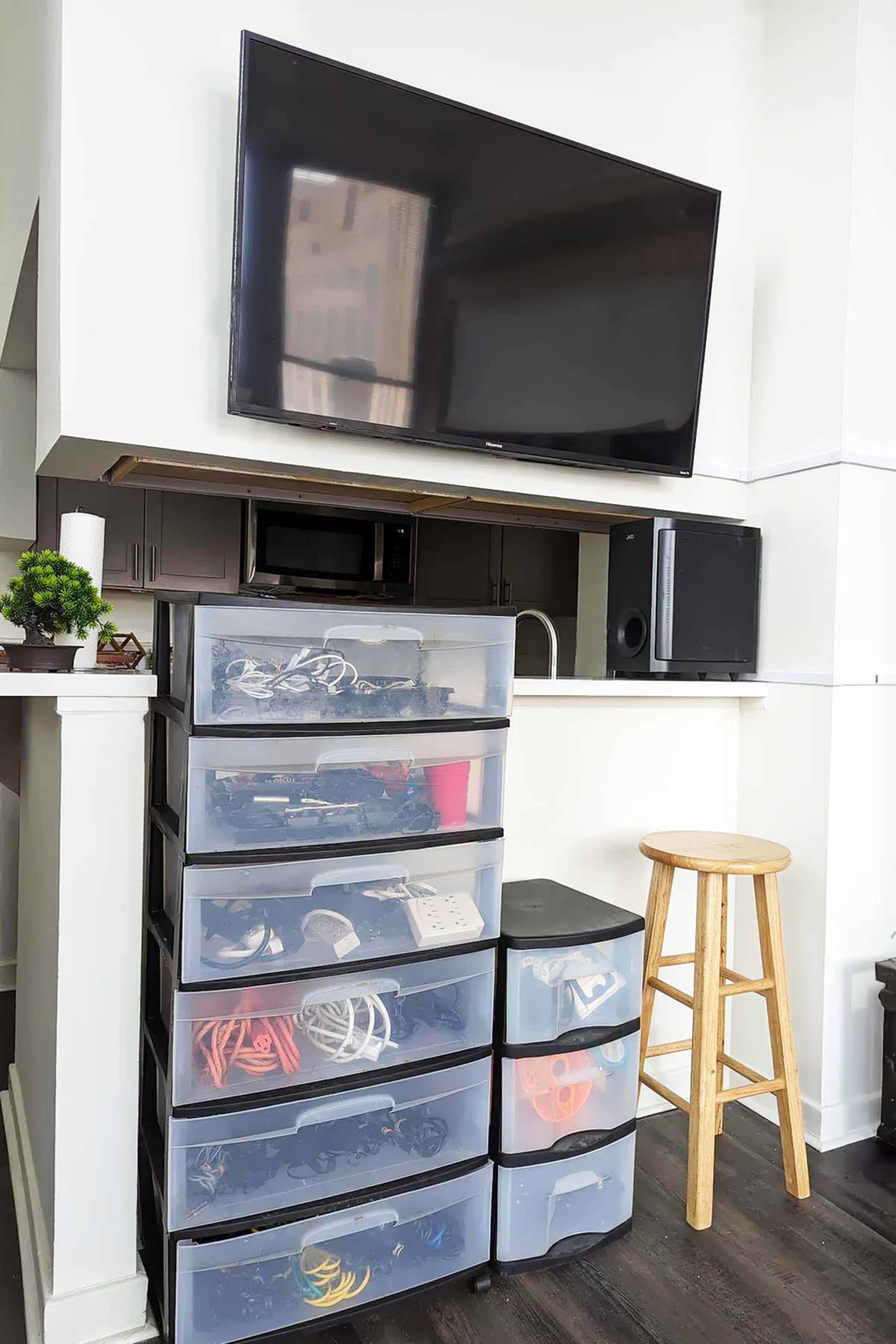 A stack of clear plastic storage drawers filled with various items is positioned next to a barstool, under a mounted television and surrounded by a kitchenette area.