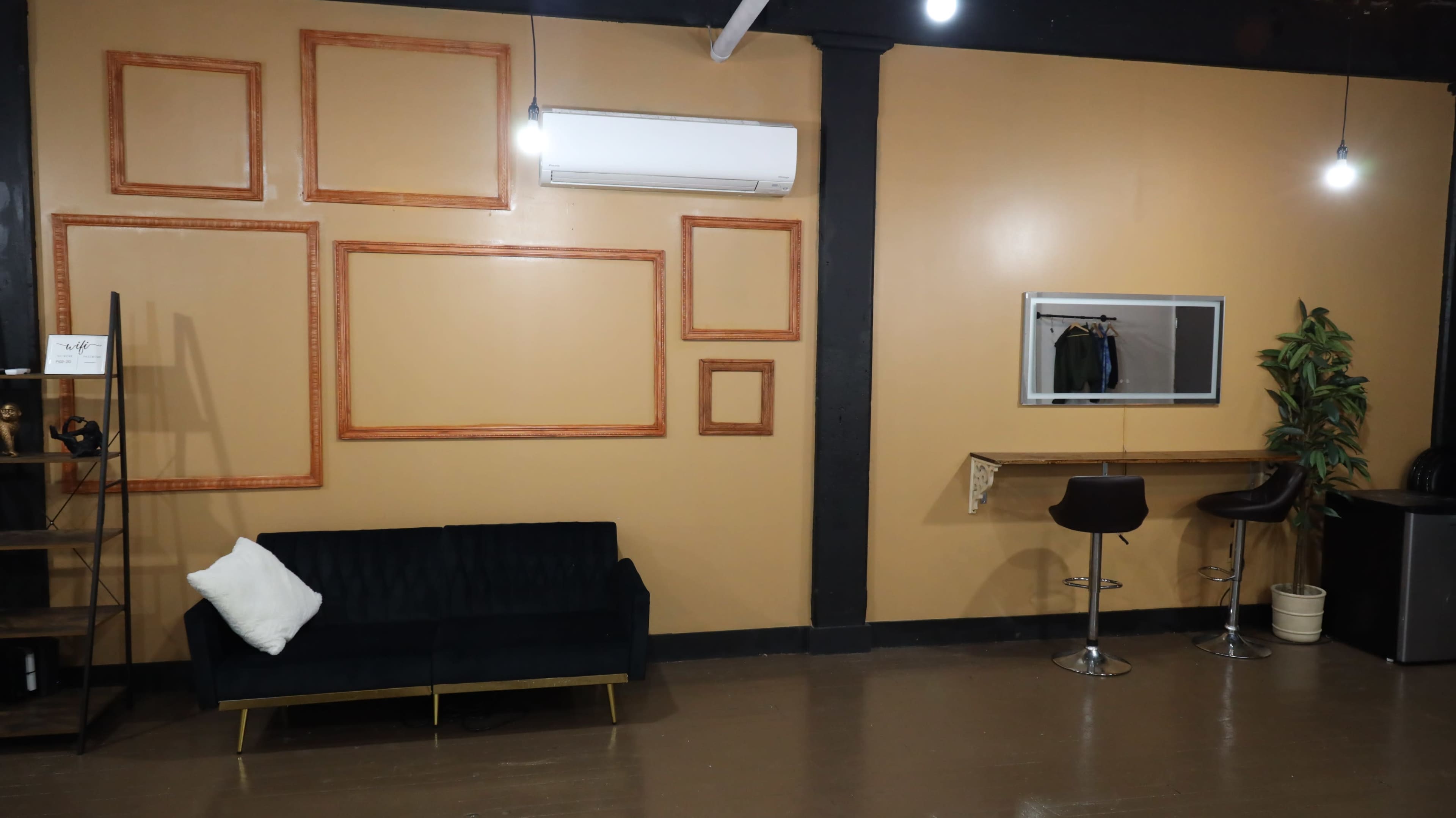 The scene features a minimalist room with a dark green sofa, decorative empty frames on the walls, a mirror, and a small bar-style table with two stools.