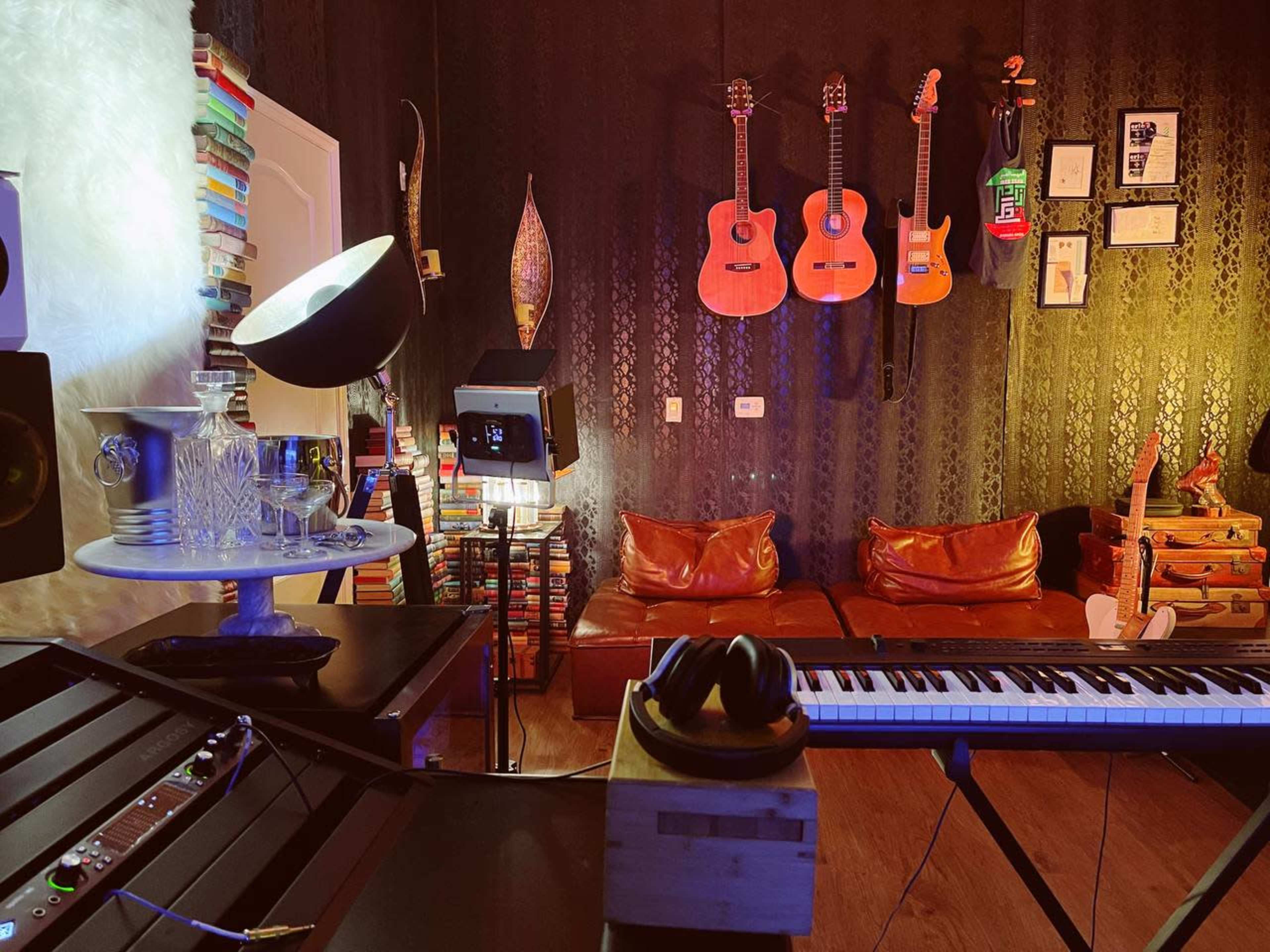 A music studio featuring guitars on the wall, a keyboard, a seating area with brown couches, and shelves filled with books.