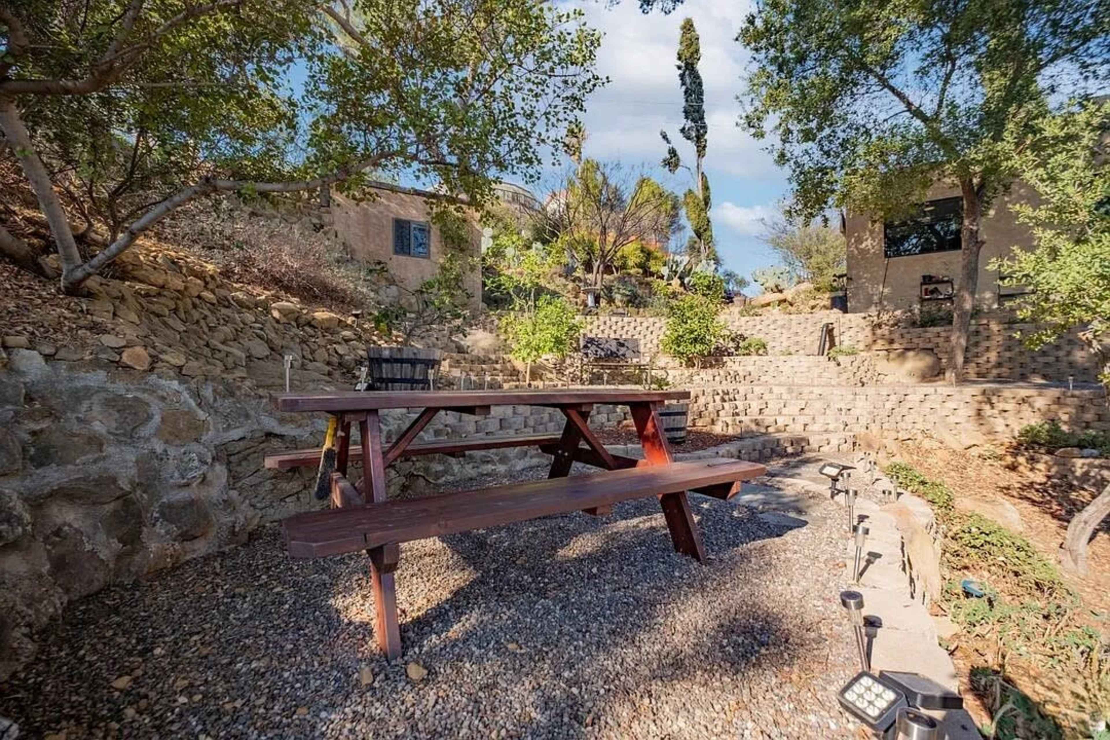 A wooden picnic table sits on gravel in a landscaped area surrounded by stone walls and trees.