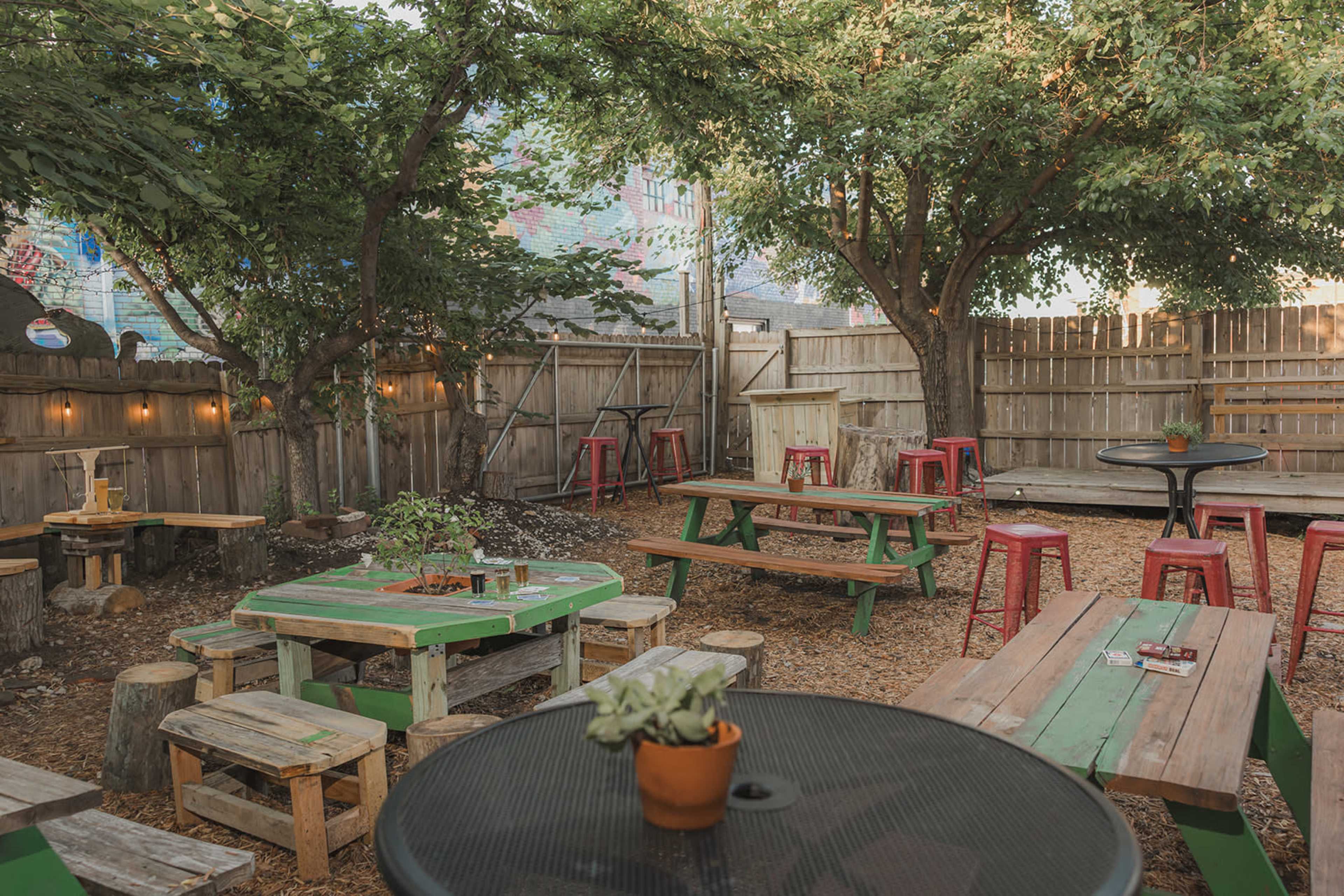 The image shows an outdoor seating area with wooden tables and benches surrounded by trees and a wooden fence.