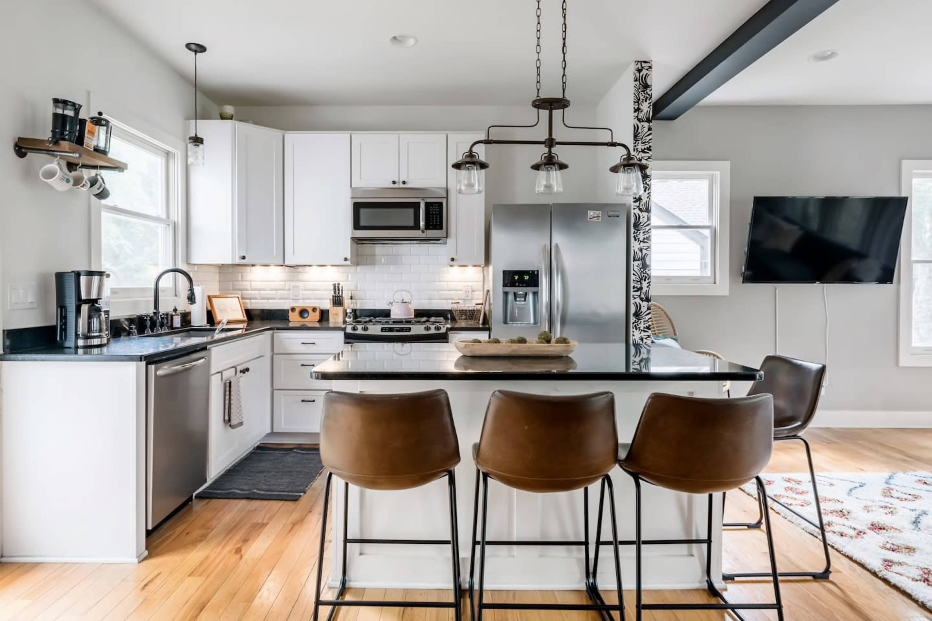 A modern kitchen with white cabinetry, stainless steel appliances, a large island with four brown bar stools, and a television mounted on the wall.