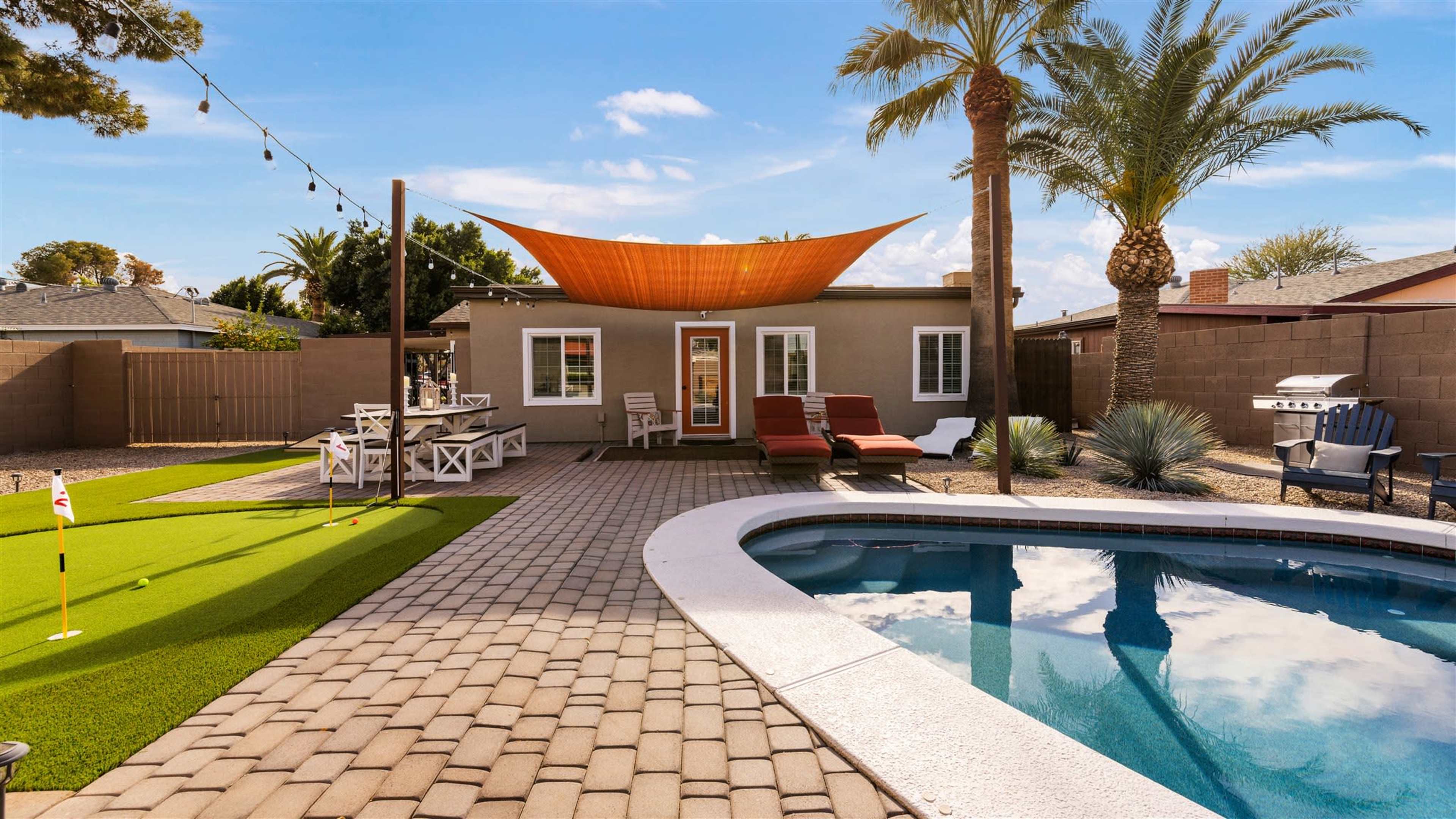 A backyard featuring a swimming pool, patio area with lounge chairs, artificial grass, and a shaded seating area, bordered by palm trees and a fence.