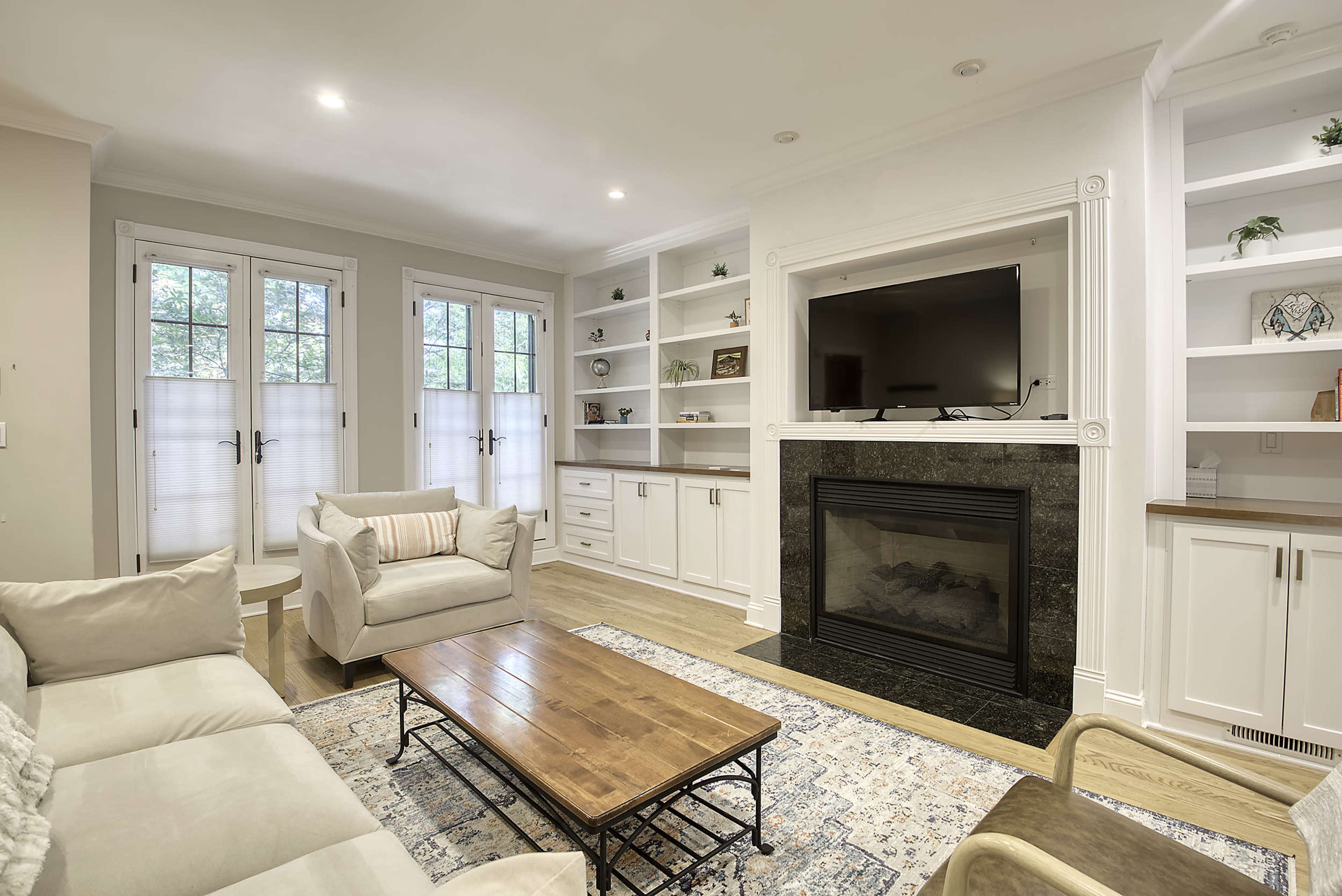 The image shows a well-lit living room featuring a beige sofa, a wooden coffee table, and a fireplace surrounded by shelves with decor and a television mounted above.