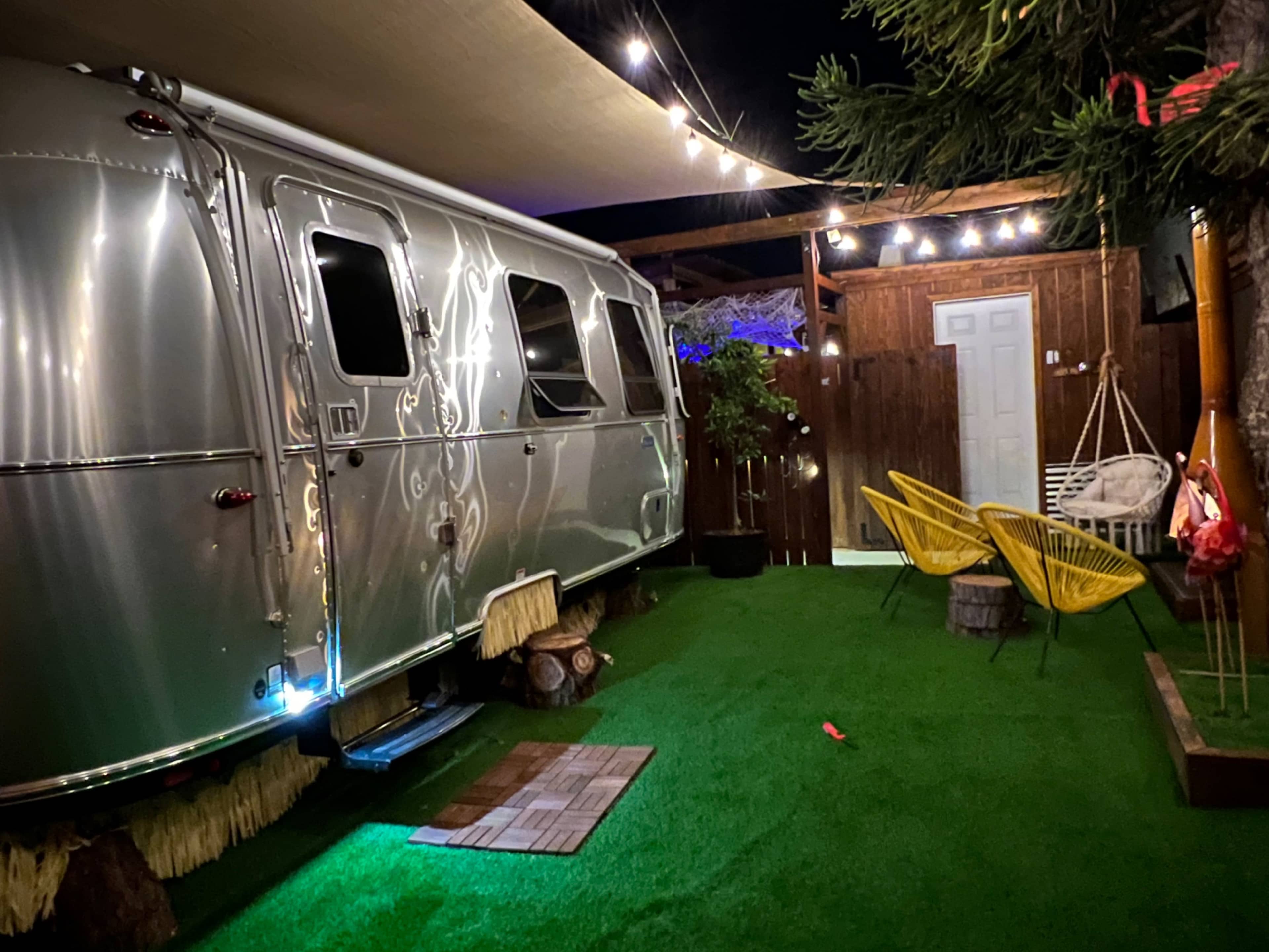 The image shows a silver Airstream trailer parked on grass surrounded by string lights, a wooden fence, and two yellow chairs.