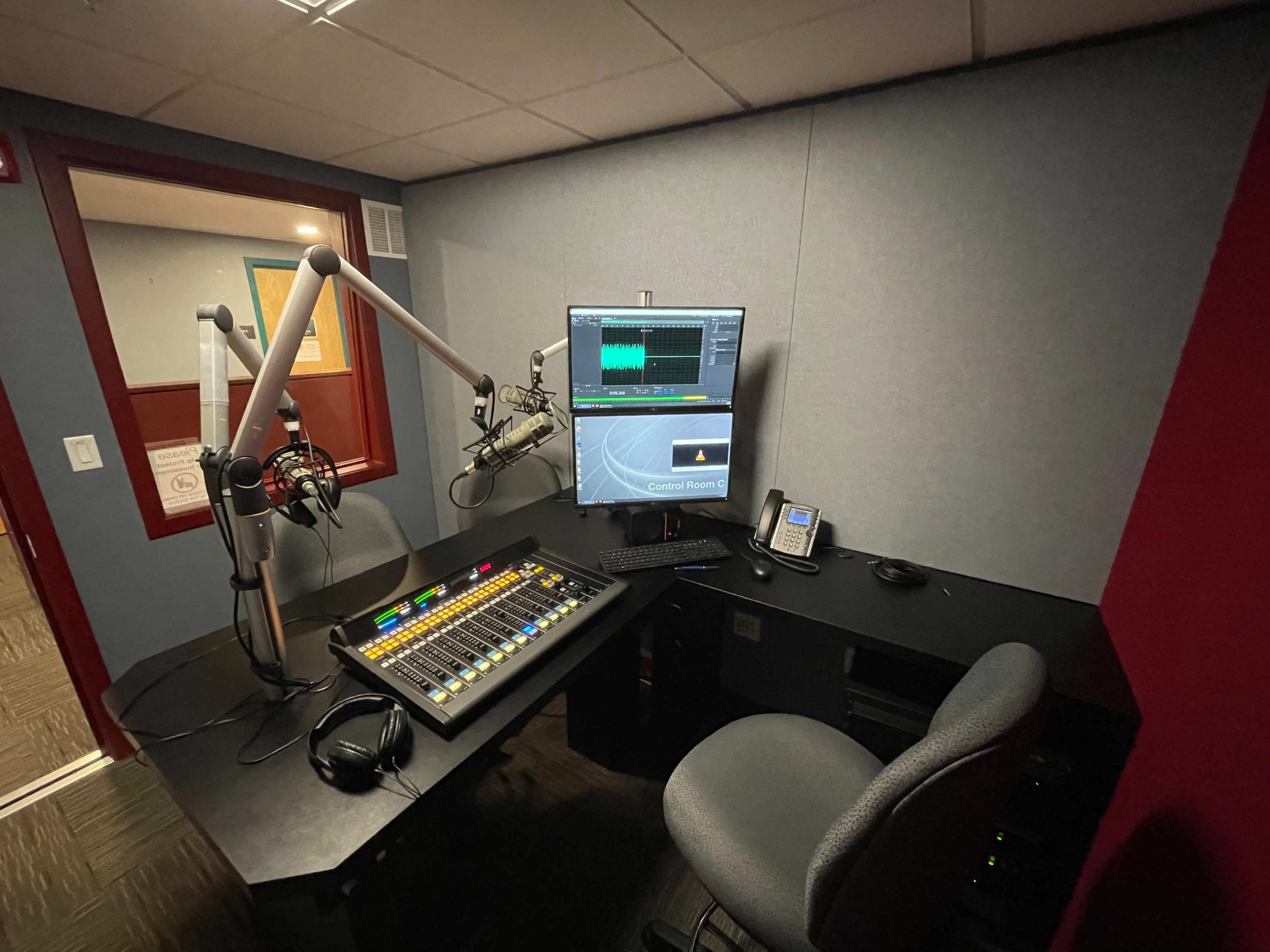 A radio studio with a control panel, microphone, computer monitor, and a chair in a small, sound-treated room.