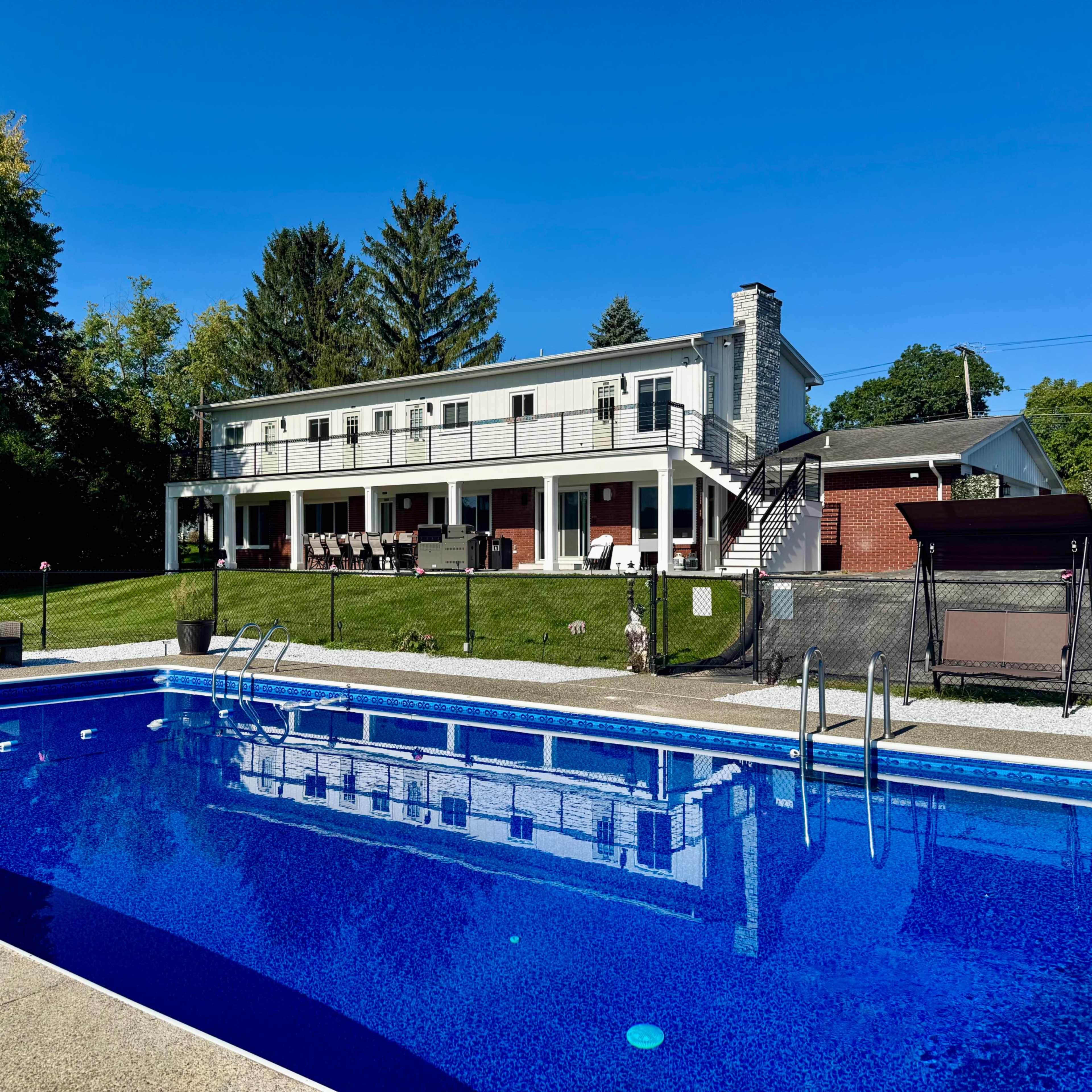A two-story house with a balcony overlooks a blue swimming pool surrounded by a grassy area and trees.