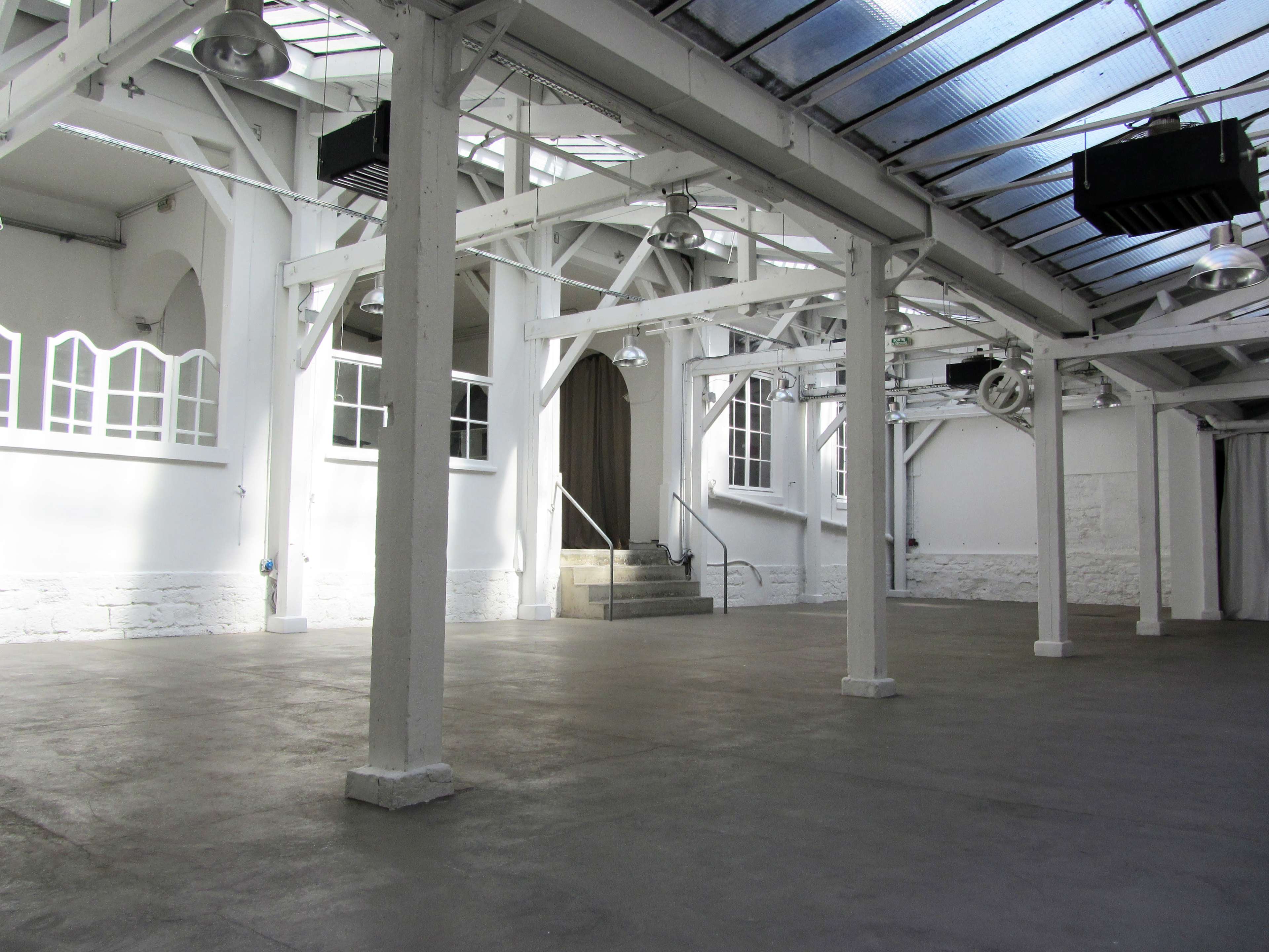 The image shows a spacious, empty industrial-style room with white walls, exposed beams, and large windows, featuring steps leading to a doorway at one end.
