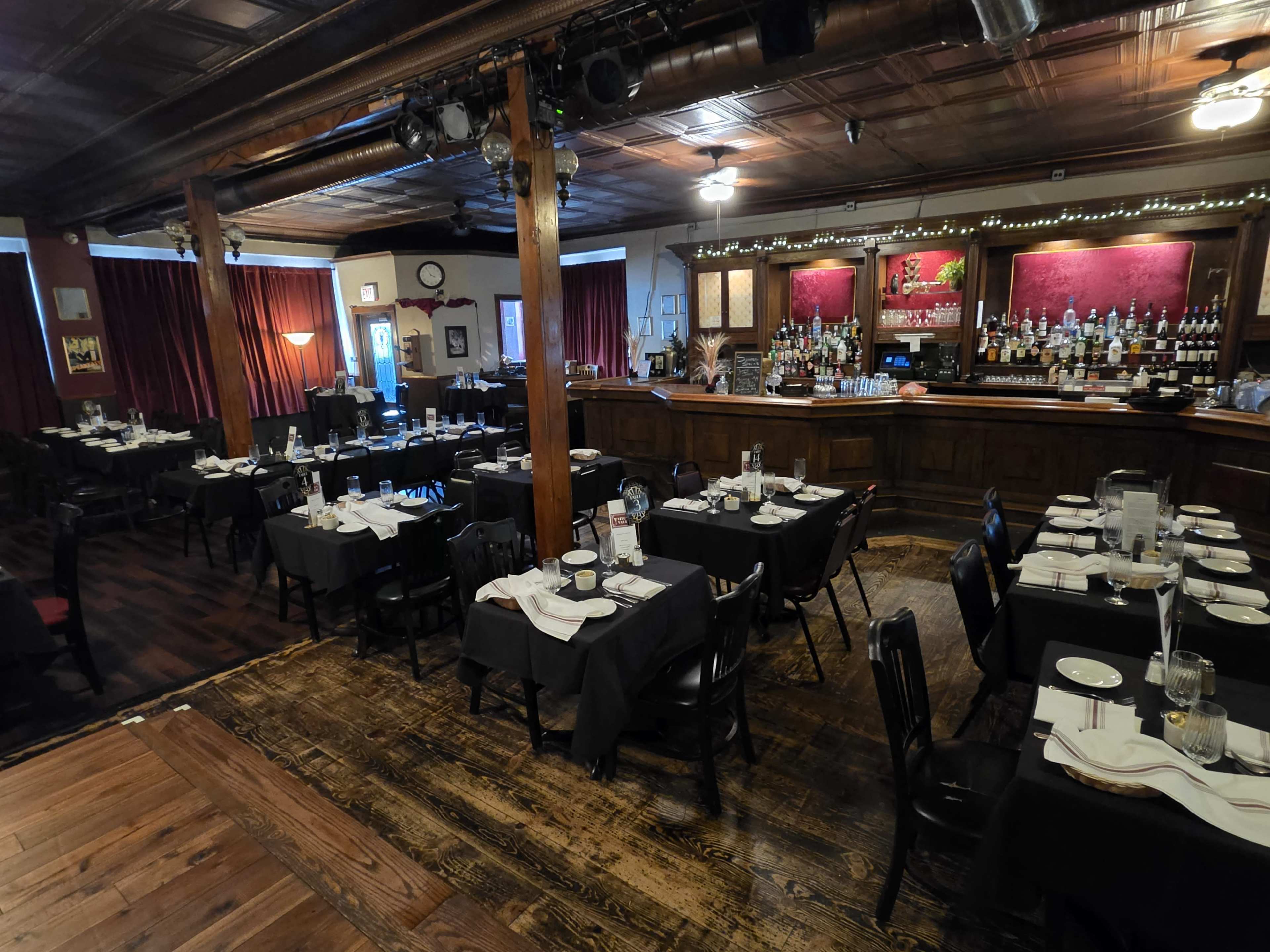 The image shows the interior of a restaurant with neatly arranged black tables and chairs, dim lighting, and a bar with a variety of bottles in the background.