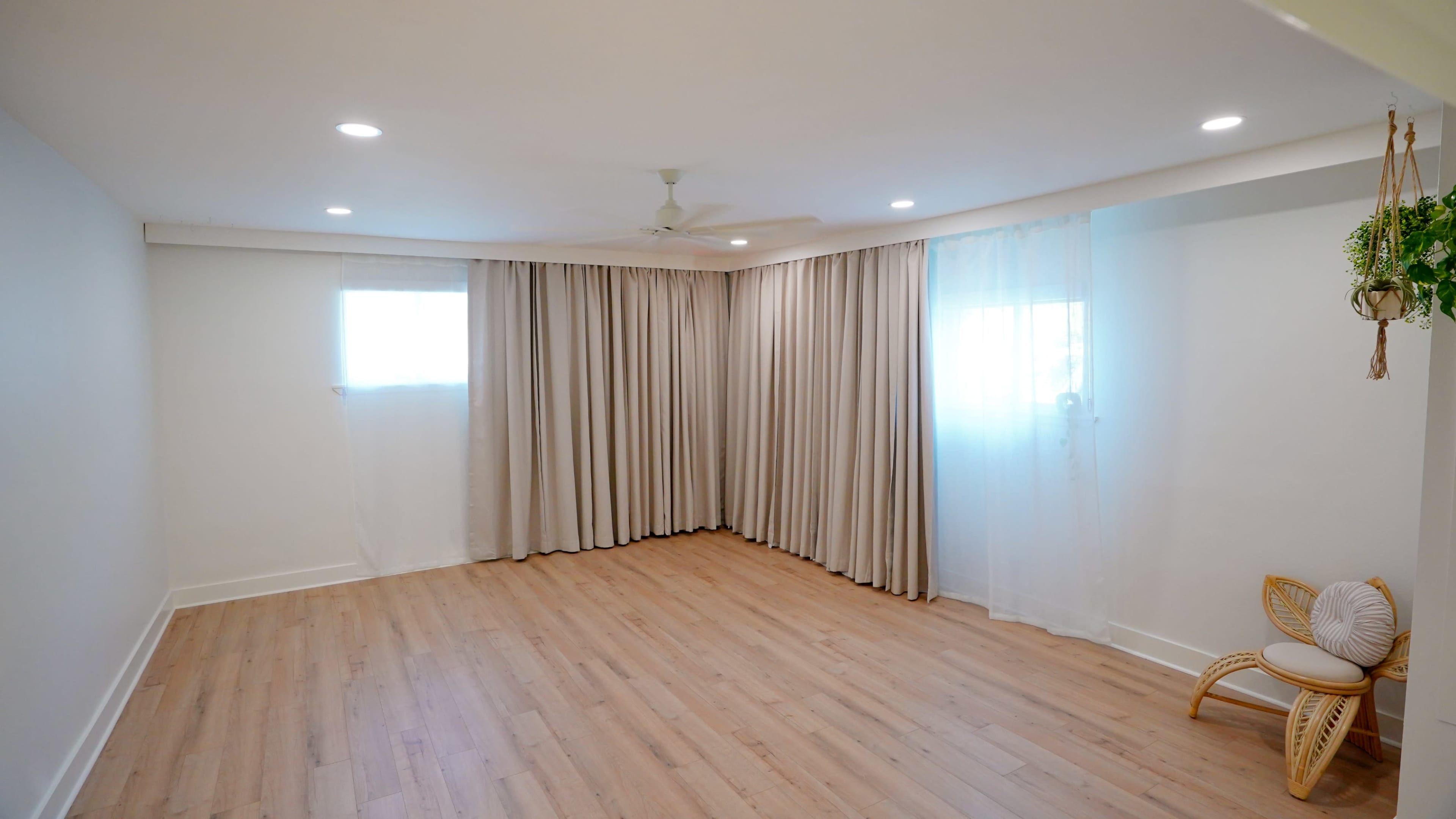 The image shows a minimally furnished room with light hardwood flooring, two windows covered by sheer and thick curtains, and a small decorative chair in the corner.