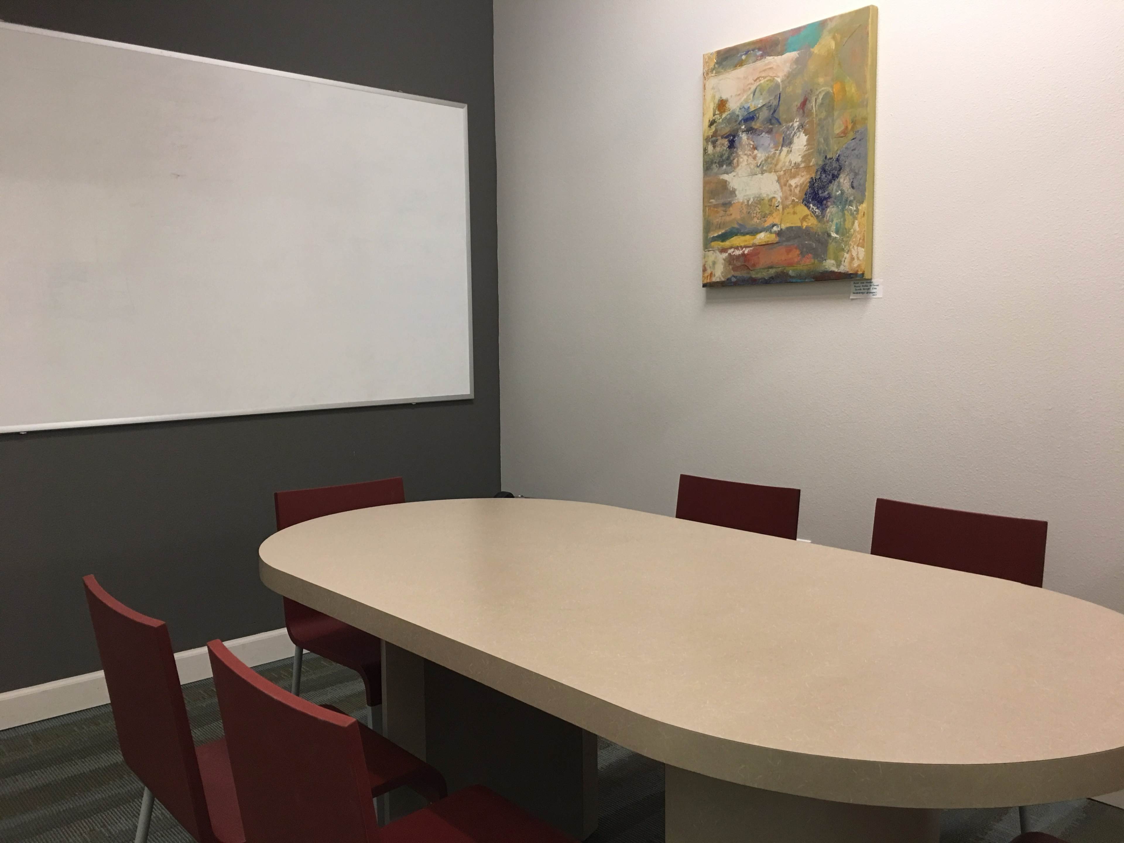 A conference room features an oval table surrounded by four red chairs, with a blank whiteboard on the wall and an abstract painting above.