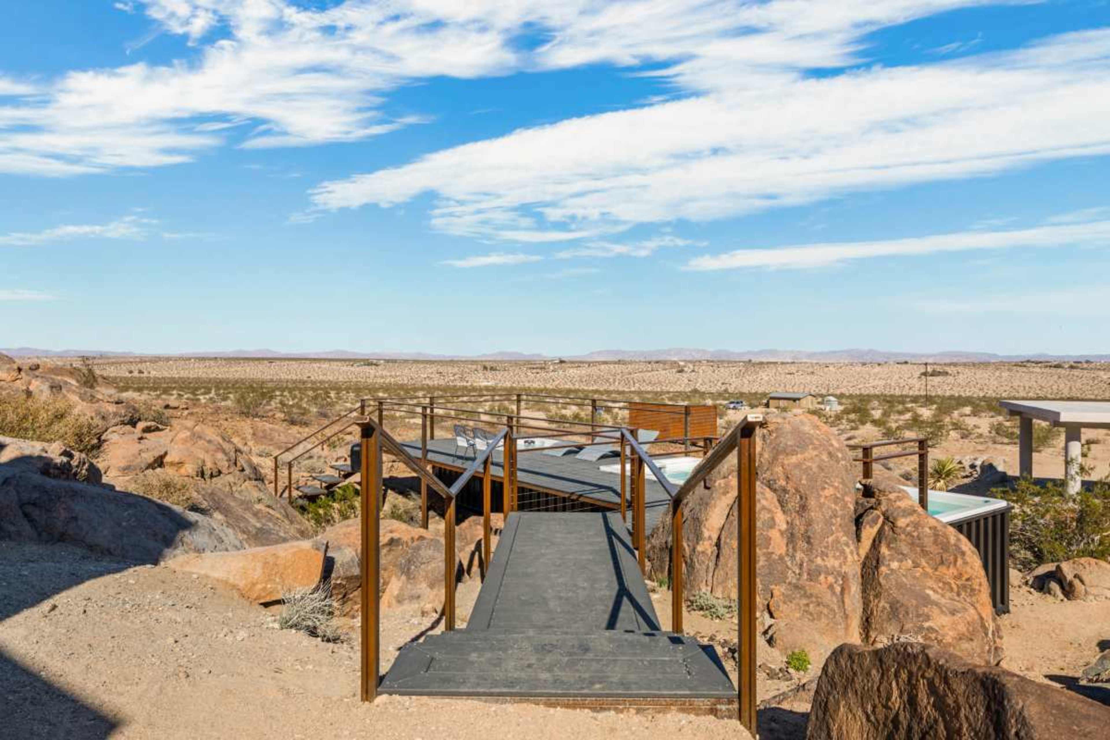The image shows a wooden walkway leading through rocky terrain, with a vast desert landscape under a blue sky in the background.