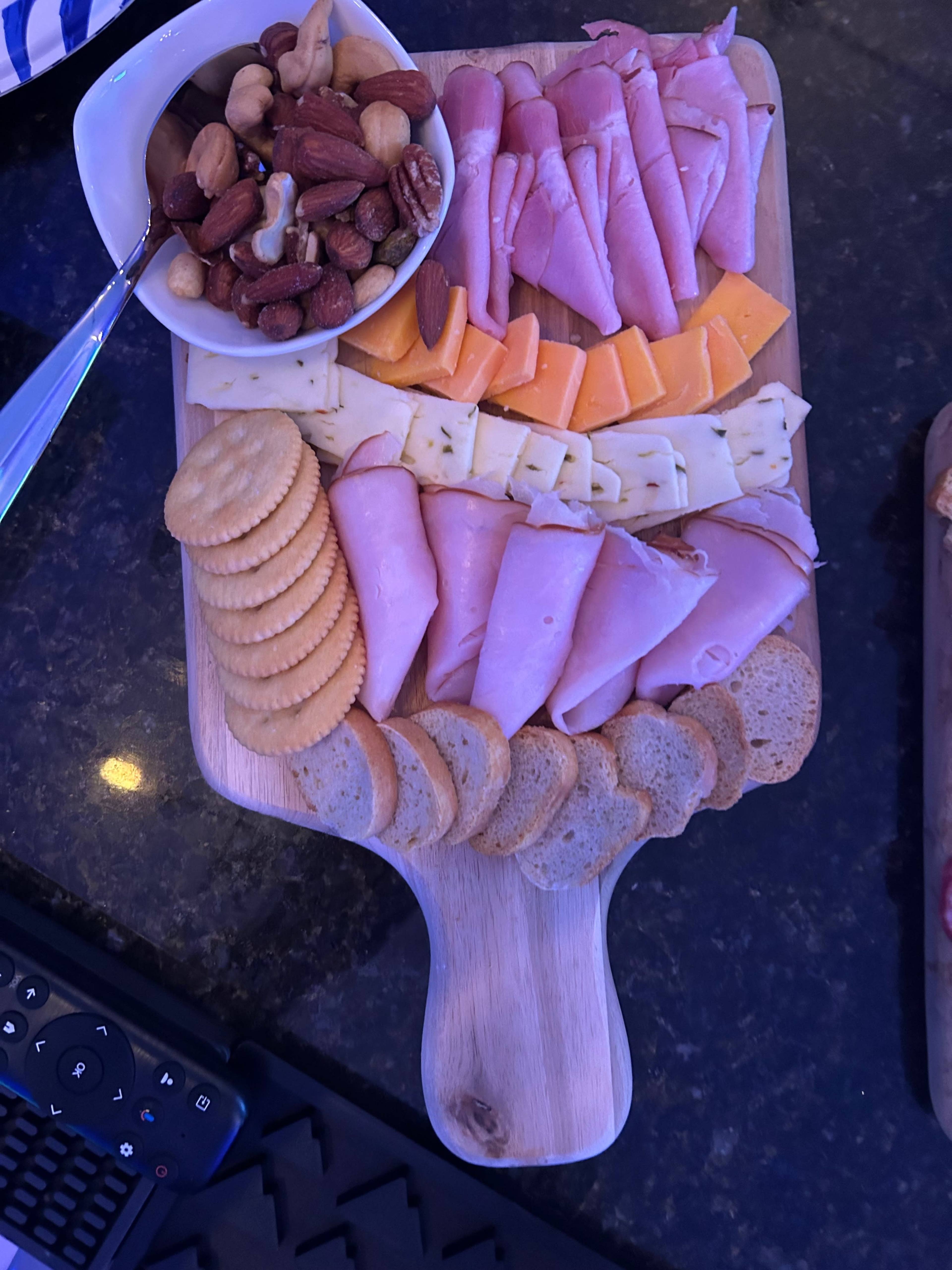 A wooden serving board displays a variety of sliced meats, cheeses, crackers, and a bowl of mixed nuts.
