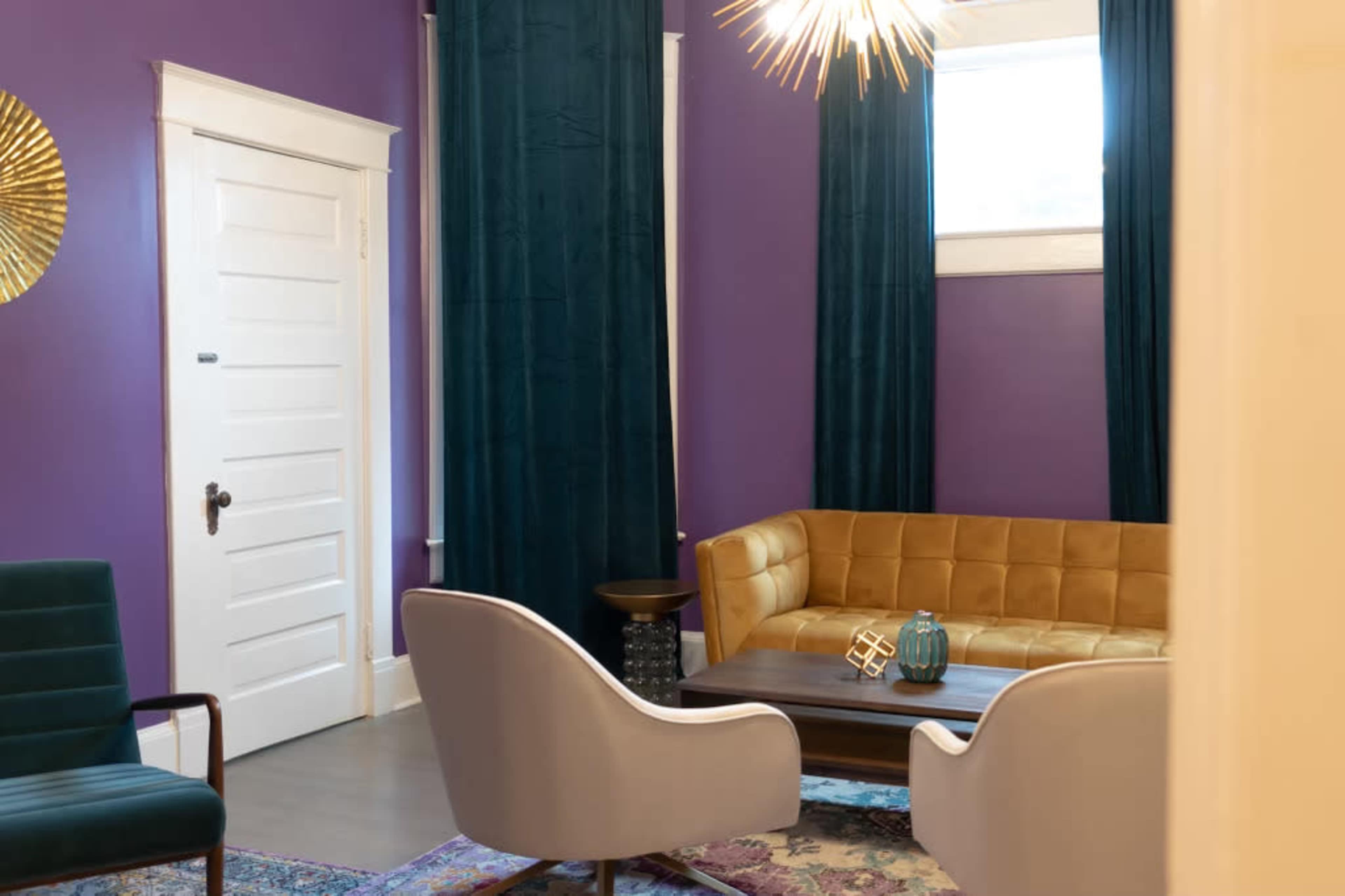 A cozy interior features a purple wall, a brown leather sofa, two gray chairs, and decorative curtains.