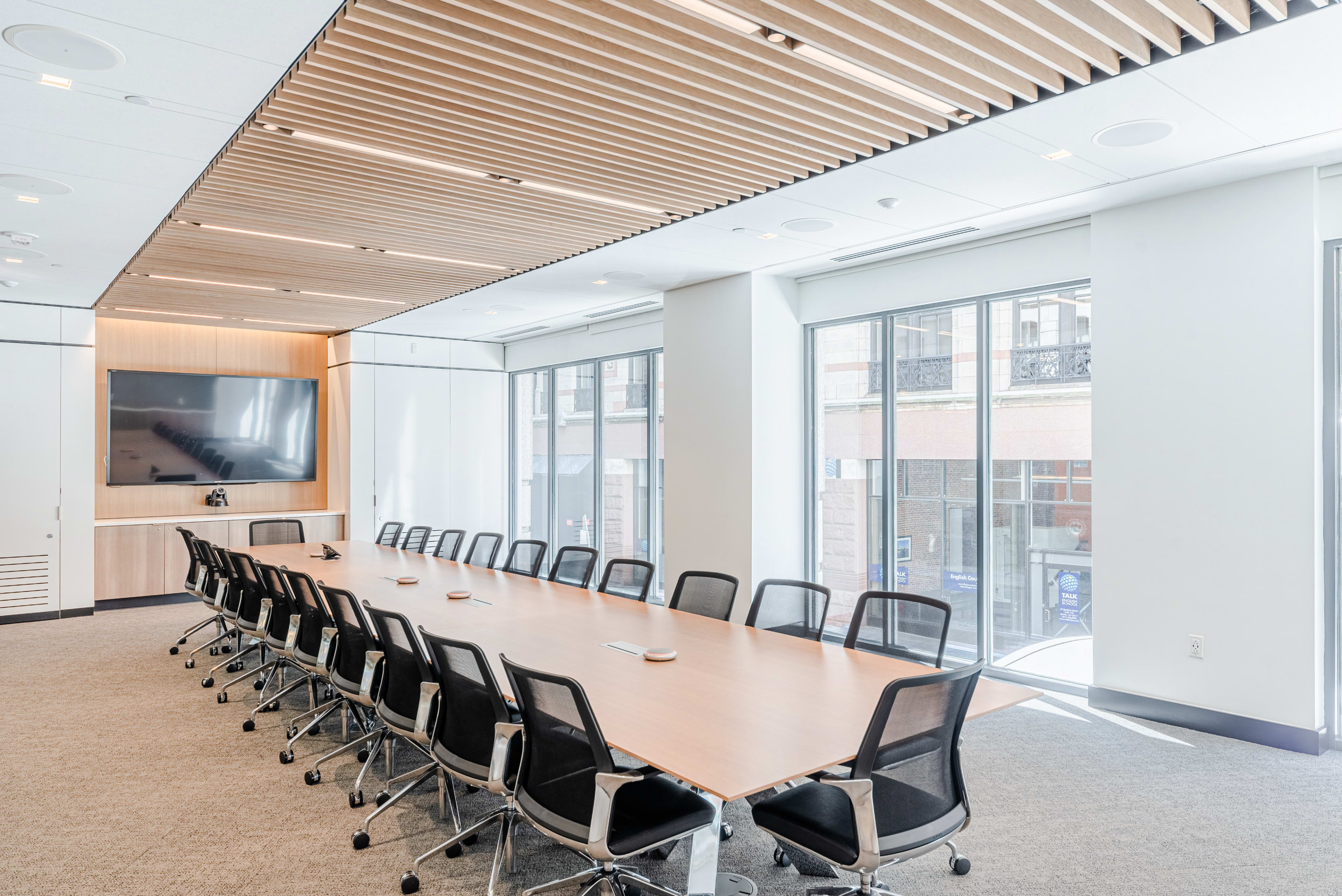 A long conference table with black chairs is set up in a modern meeting room featuring large windows and a wall-mounted screen.