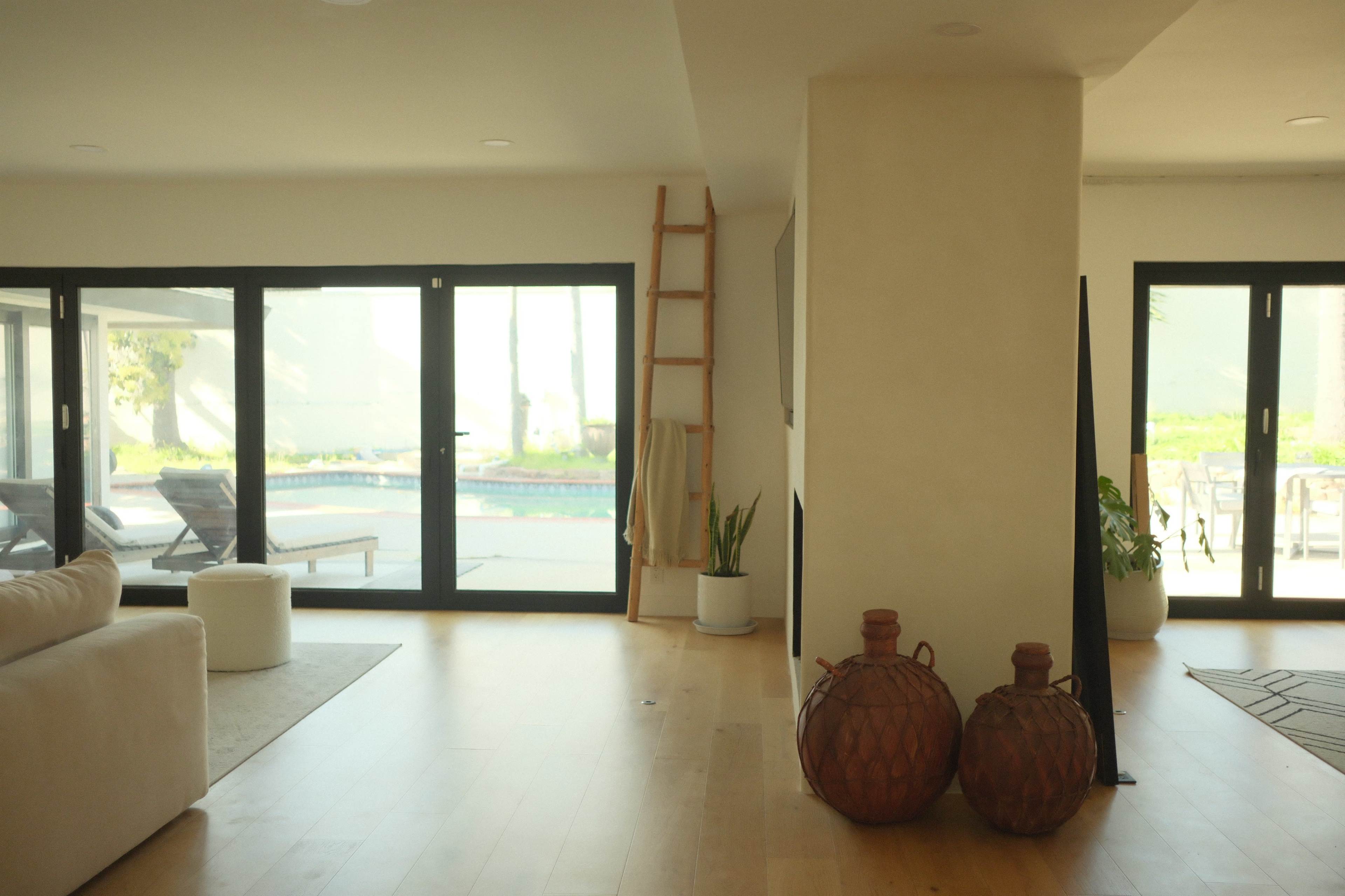 The image shows a modern interior space featuring a sofa, a decorative ladder, and clay pots, with large glass doors leading to an outdoor pool area.
