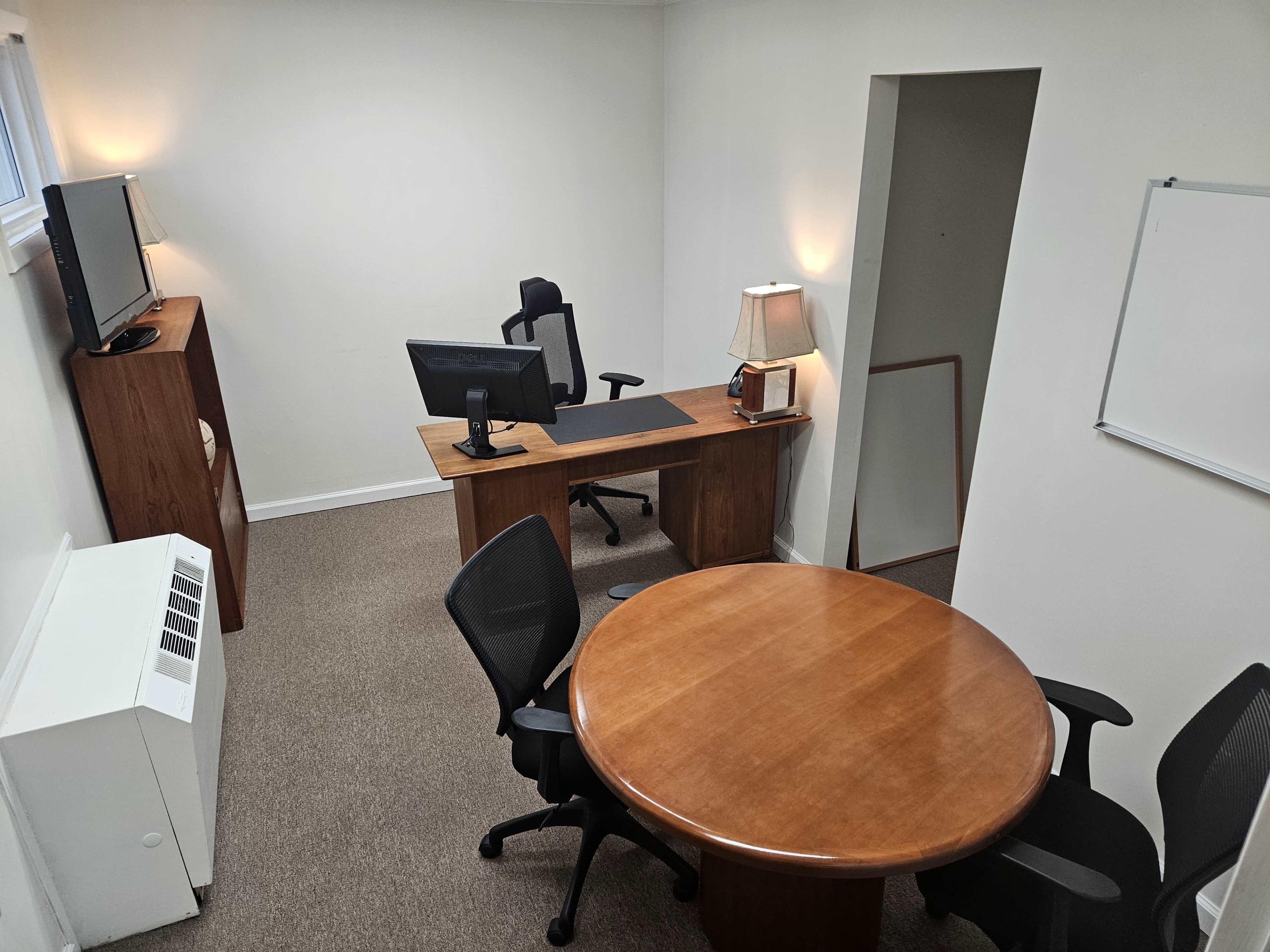 The image shows a small office space with a desk, a round table, and a television on a stand.