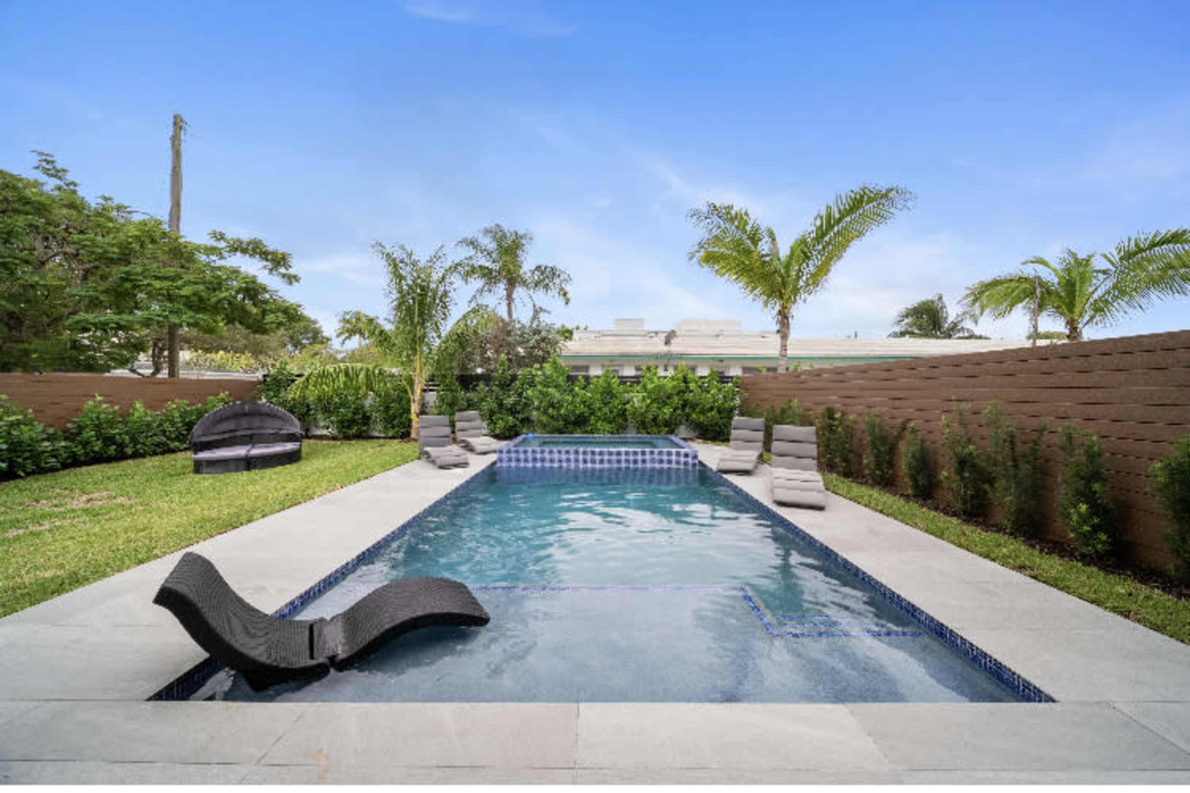 The image shows a rectangular swimming pool surrounded by lounge chairs and palm trees, enclosed by a low wall and greenery.