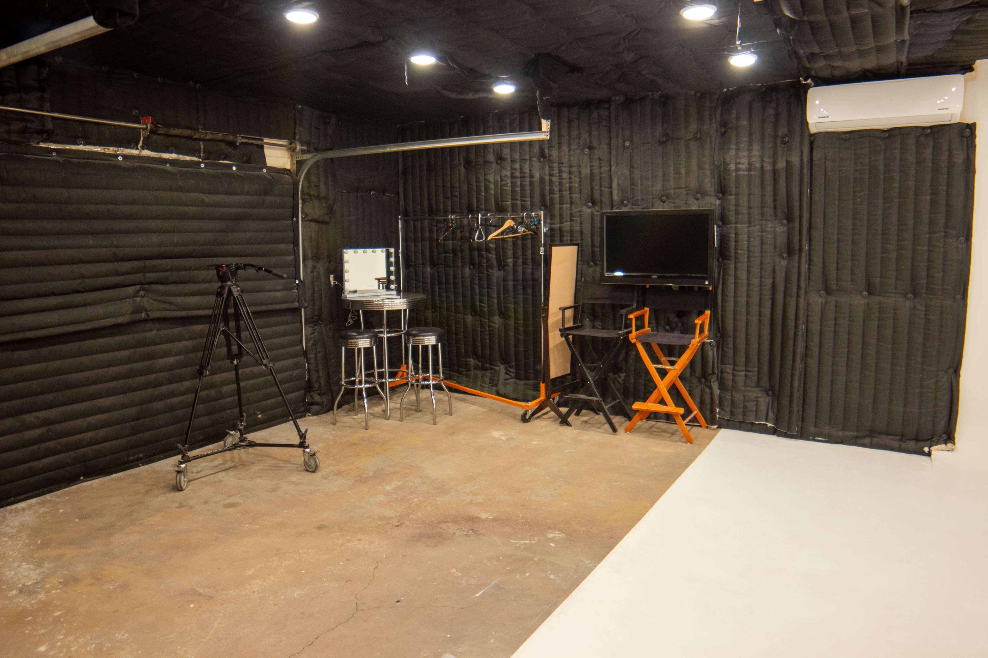 The image shows a studio space with black padded walls, a monitor on an orange frame, two tall stools, a clothing rack, and a tripod positioned in the corner.