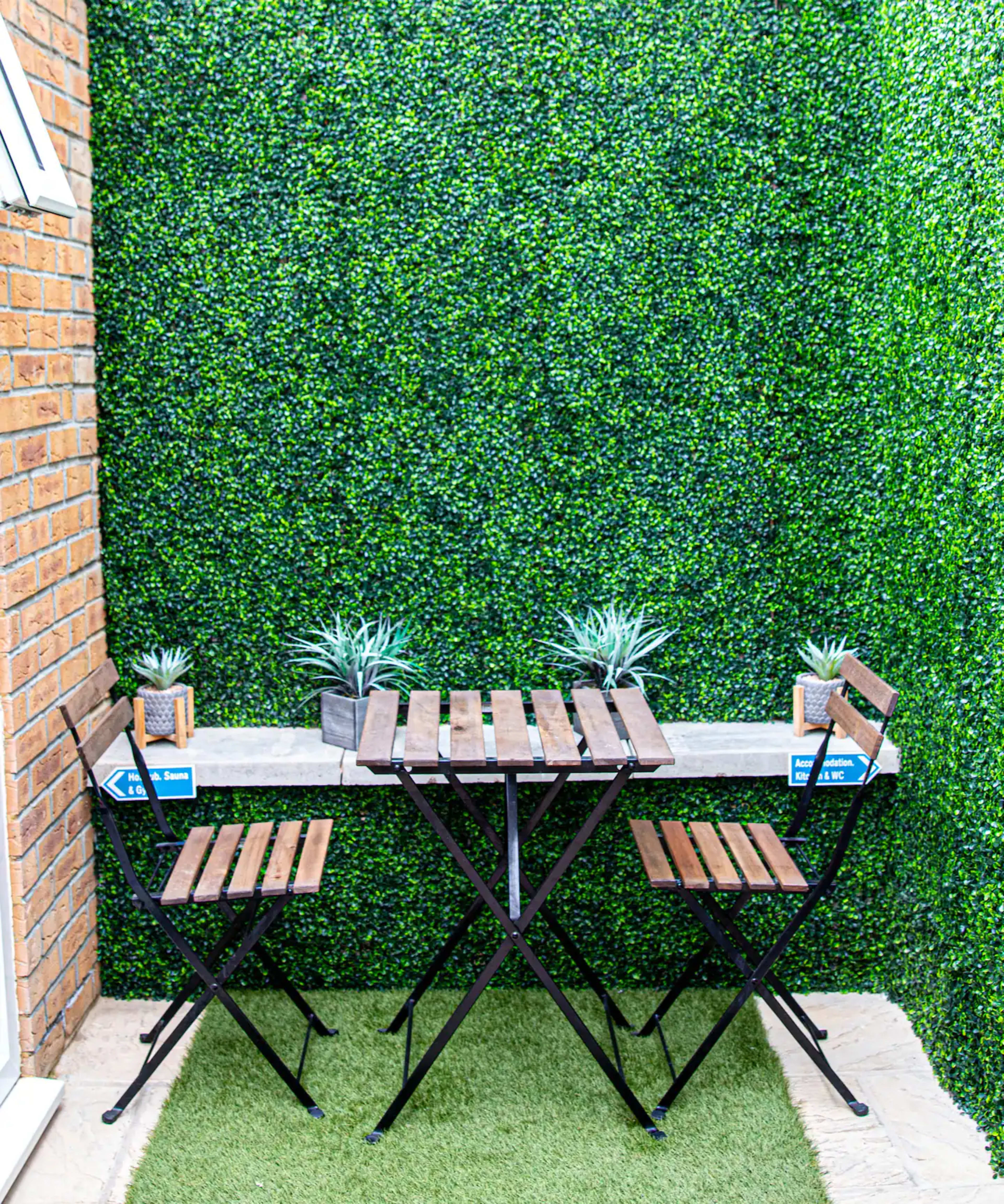 A small outdoor seating area features a wooden table with two folding chairs and potted plants against a green wall of artificial foliage.