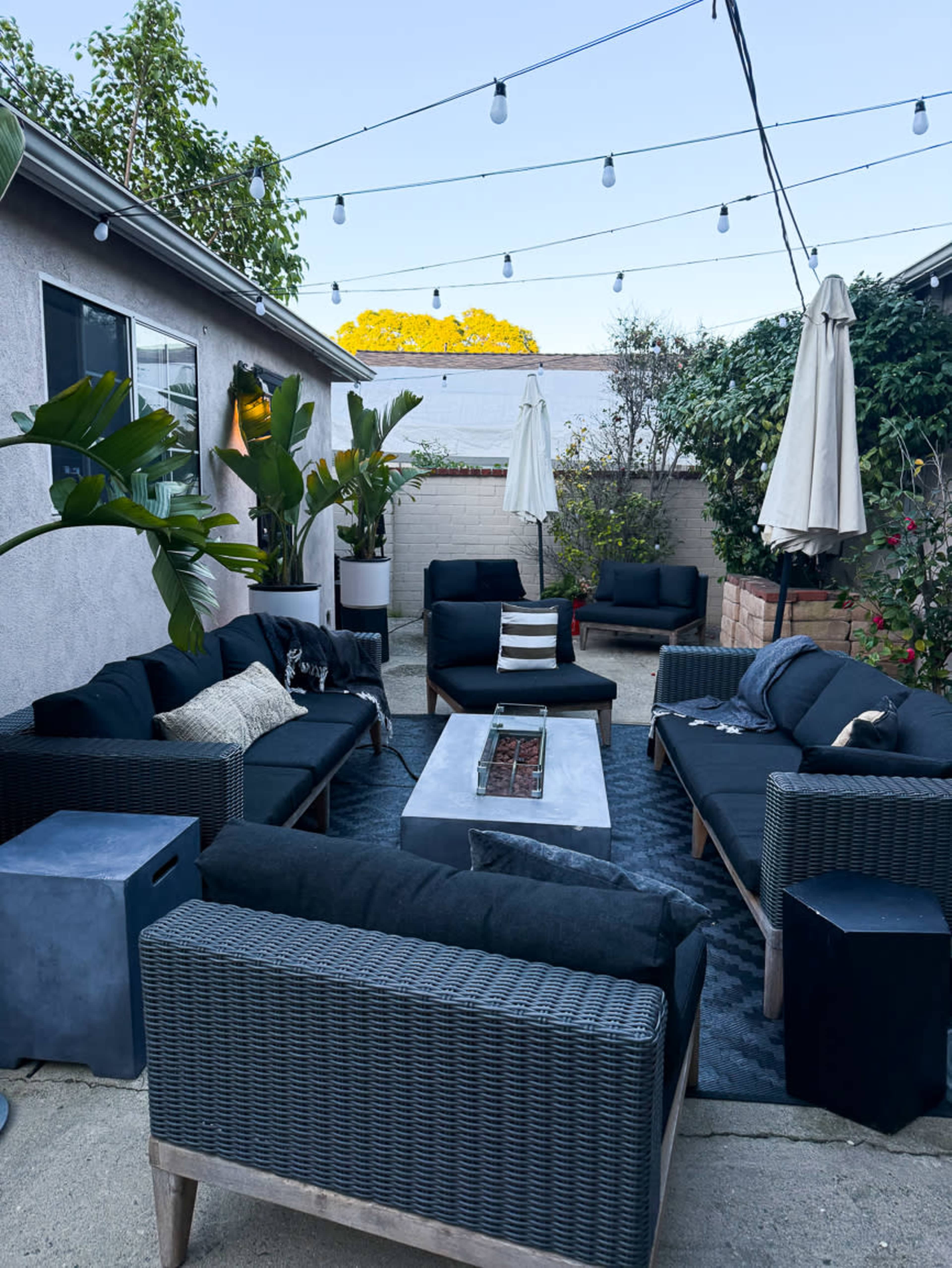 The image shows a landscaped outdoor seating area with black wicker furniture arranged around a central fire pit, surrounded by plants and string lights overhead.