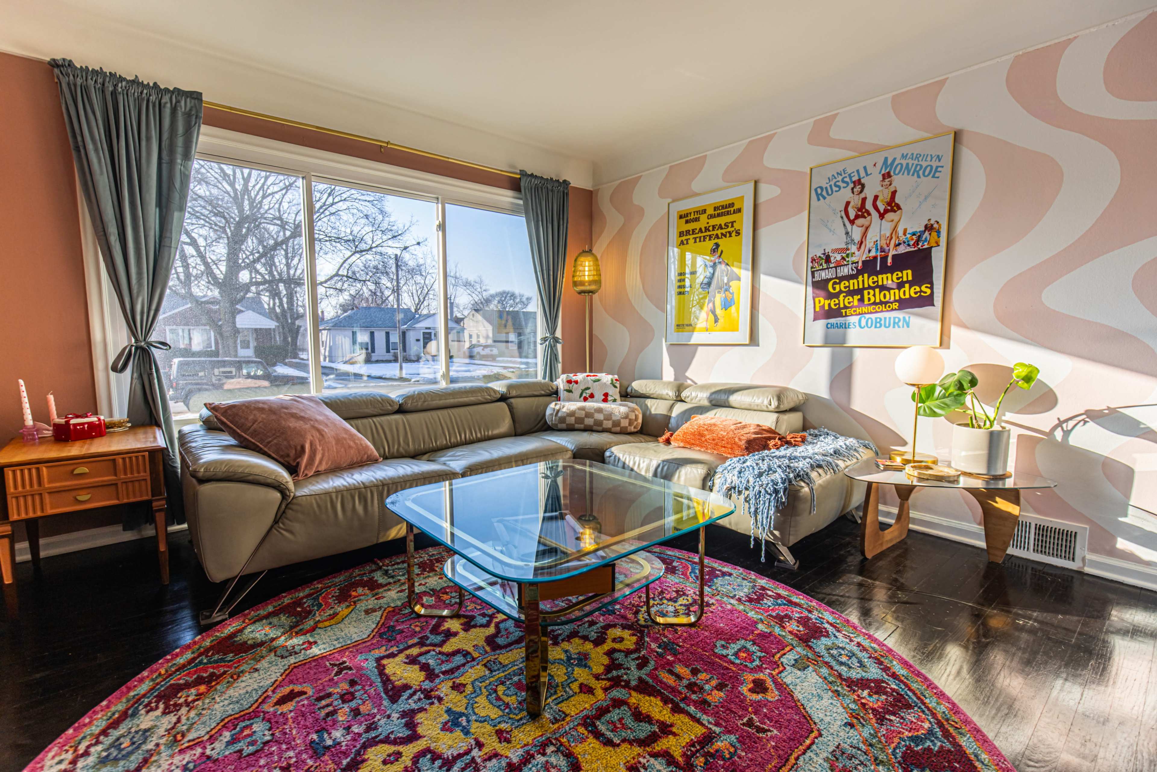A stylish living room features a sectional sofa, a glass coffee table, and colorful artwork on the walls, illuminated by natural light from a large window.